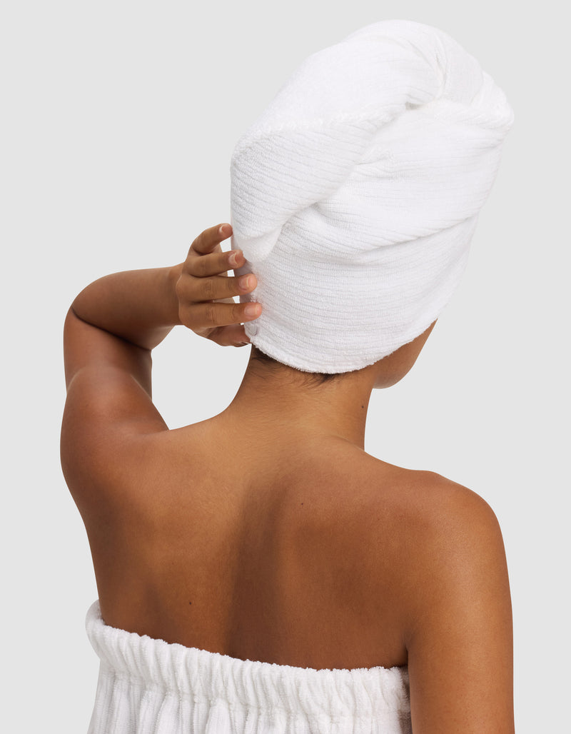 A person with medium skin tone is seen from behind, wearing a white towel around their body and holding a Cozy Earth Ribbed Terry Hair Towel wrapped around their head. The background is plain light gray.