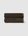 Two neatly rolled, dark brown towels from the Cozy Earth Complete Ribbed Terry Bath Bundle are stacked against a plain light gray background.|Color:Espresso