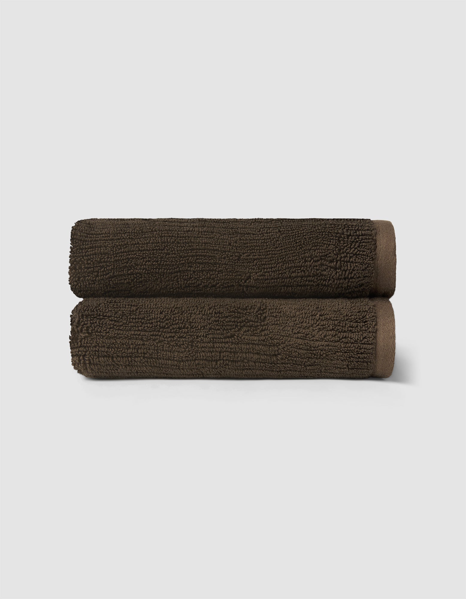 Two neatly rolled, dark brown towels from the Cozy Earth Complete Ribbed Terry Bath Bundle are stacked against a plain light gray background.|Color:Espresso