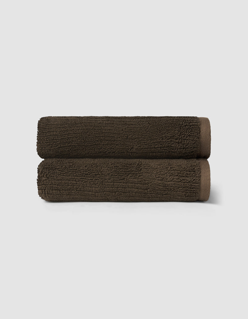 Two neatly rolled, dark brown towels from the Cozy Earth Complete Ribbed Terry Bath Bundle are stacked against a plain light gray background.
