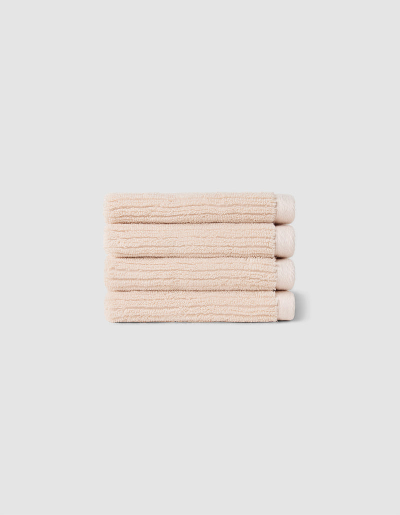 The Complete Ribbed Terry Bath Bundle by Cozy Earth features four neatly folded beige towels stacked atop each other against a plain light gray background.