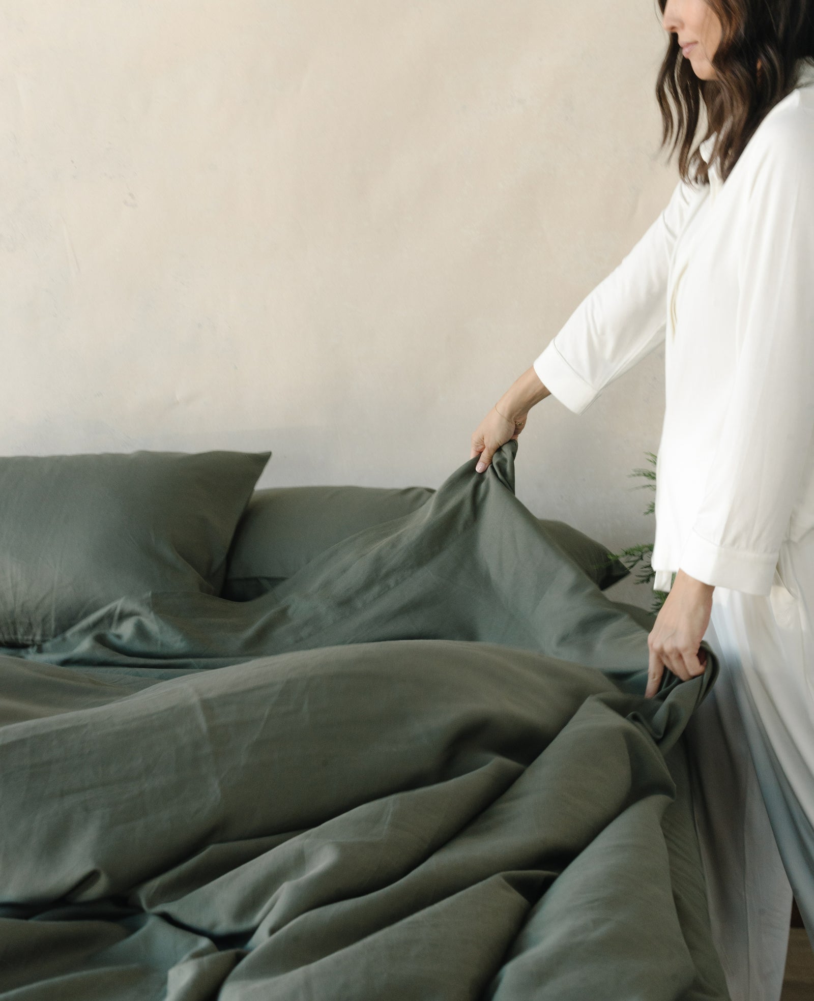 A woman in white pajamas makes a bed with dark green Cozy Earth Bamboo Sheet Set from Overstock, gently pulling the blanket up over the mattress in a softly lit, neutral-toned room. 