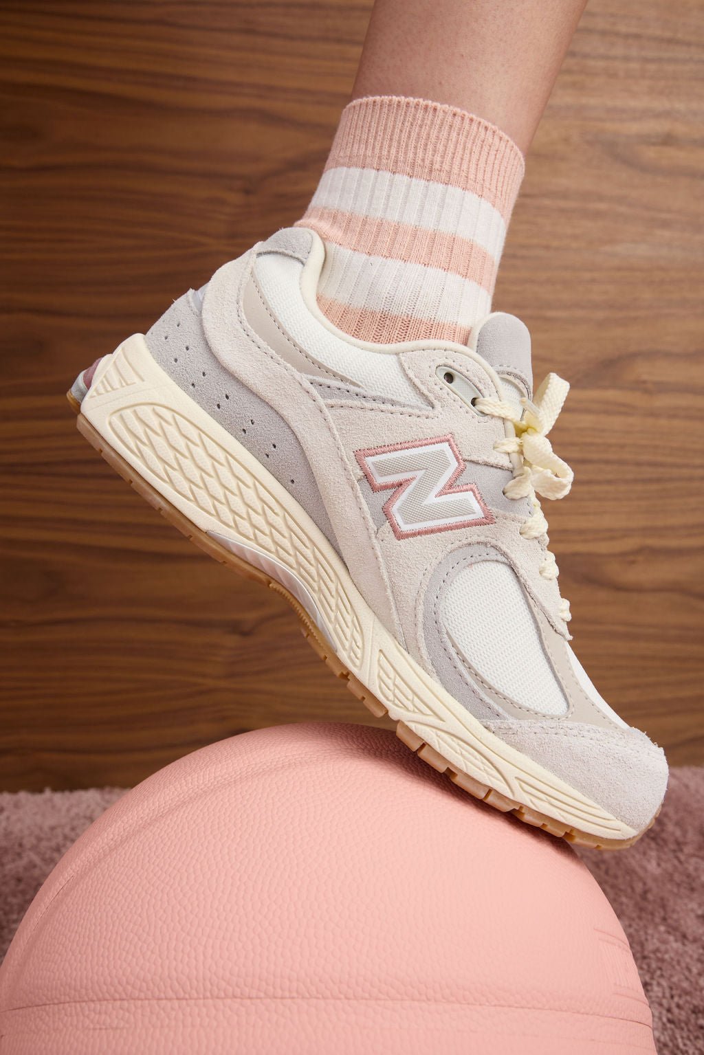 A person models the Women's Sadie Sock by Cozy Earth, featuring a peach shade with two white stripes, wearing sneakers and standing on a pink ball. 