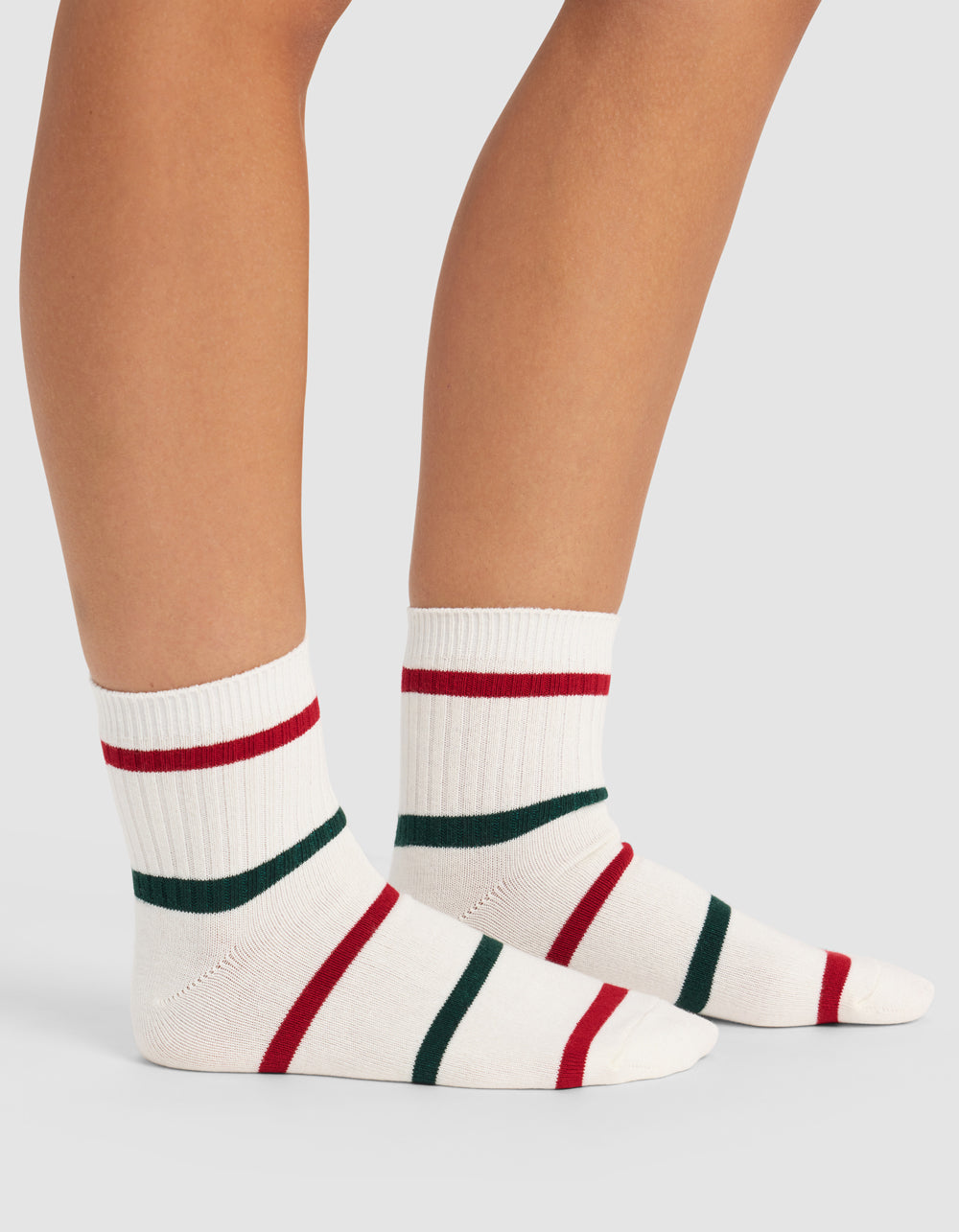 A person wears Cozy Earth Women's Sadie Socks—white ribbed crew socks with red and green stripes—standing on a light gray surface, showing only their lower legs and feet.