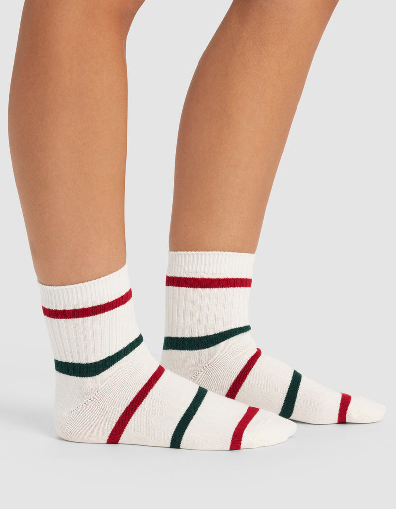 A person wears Cozy Earth Women's Sadie Socks—white ribbed crew socks with red and green stripes—standing on a light gray surface, showing only their lower legs and feet.