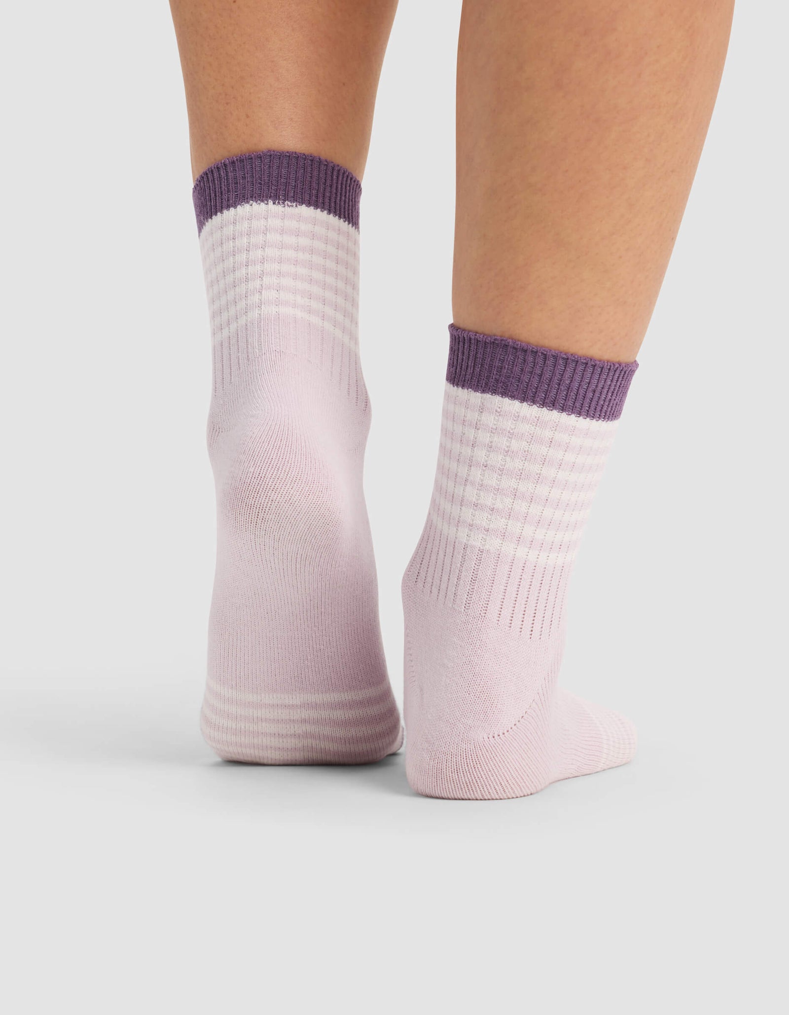 A close-up of two feet wearing Cozy Earth Women's Sadie Socks—light pink with ribbed texture and thick purple cuffs—standing on a plain light gray background.