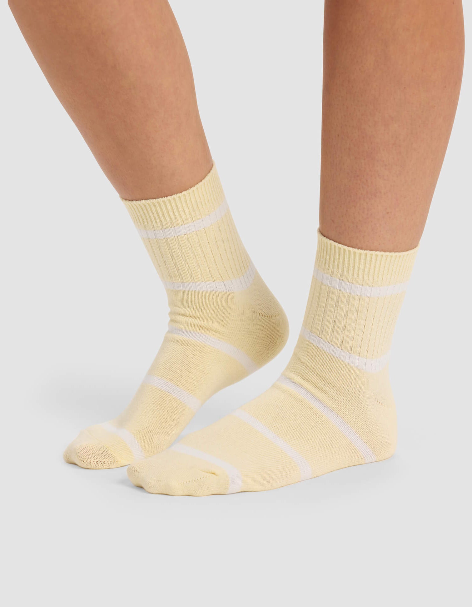A person wears Cozy Earth Women's Sadie Sock—light yellow crew socks with white stripes—standing on a light gray surface, showing only their lower legs and feet.