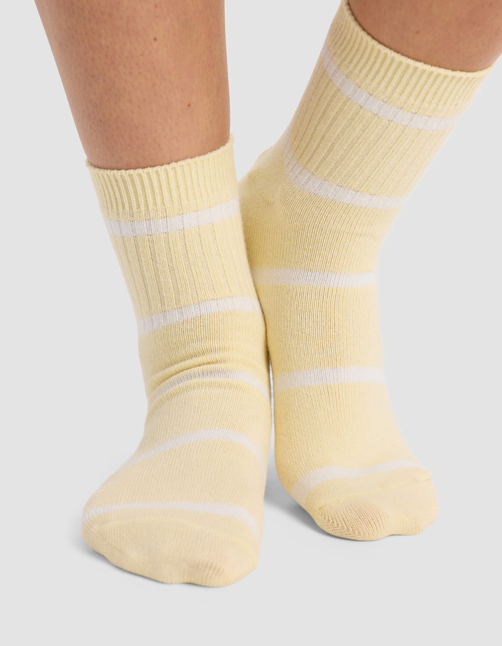 A person stands on a light gray surface wearing Cozy Earth Women's Sadie Socks in light yellow with white horizontal stripes.