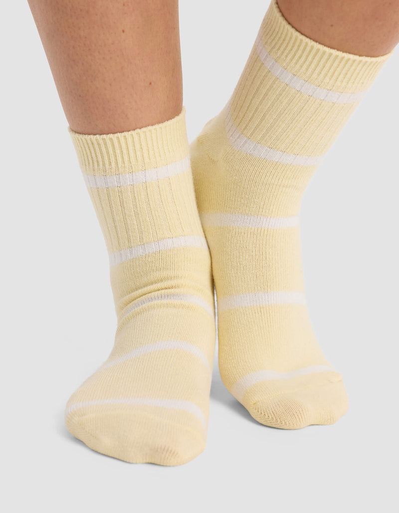 A person stands on a light gray surface wearing Cozy Earth Women's Sadie Socks in light yellow with white horizontal stripes.