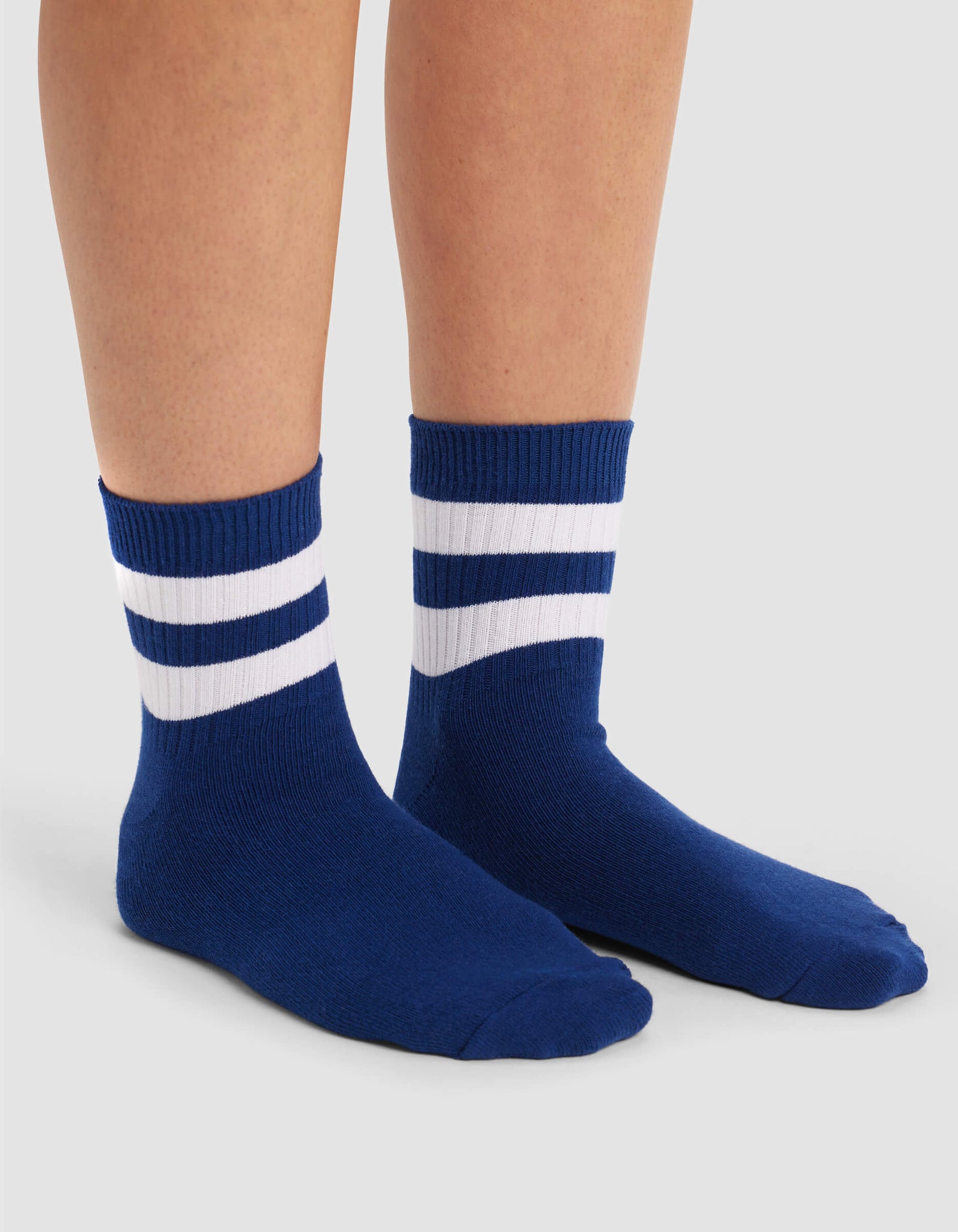 A person wears Cozy Earth's Women's Sadie Sock—blue crew socks with two white stripes near the top—while standing on a light gray background.