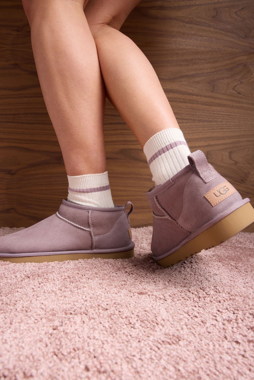 Someone wearing Cozy Earth's Women's Sadie Sock in white and pink UGG ankle boots is standing on a textured pink carpet with crossed legs against a wooden wall panel backdrop.