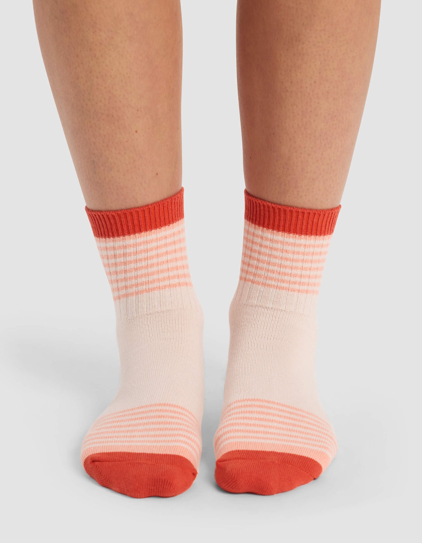 A pair of feet wear Cozy Earth's Women's Sadie Socks in light pink with red toes, heels, and cuffs, along with a subtle striped pattern near the top and bottom on a plain light background.
