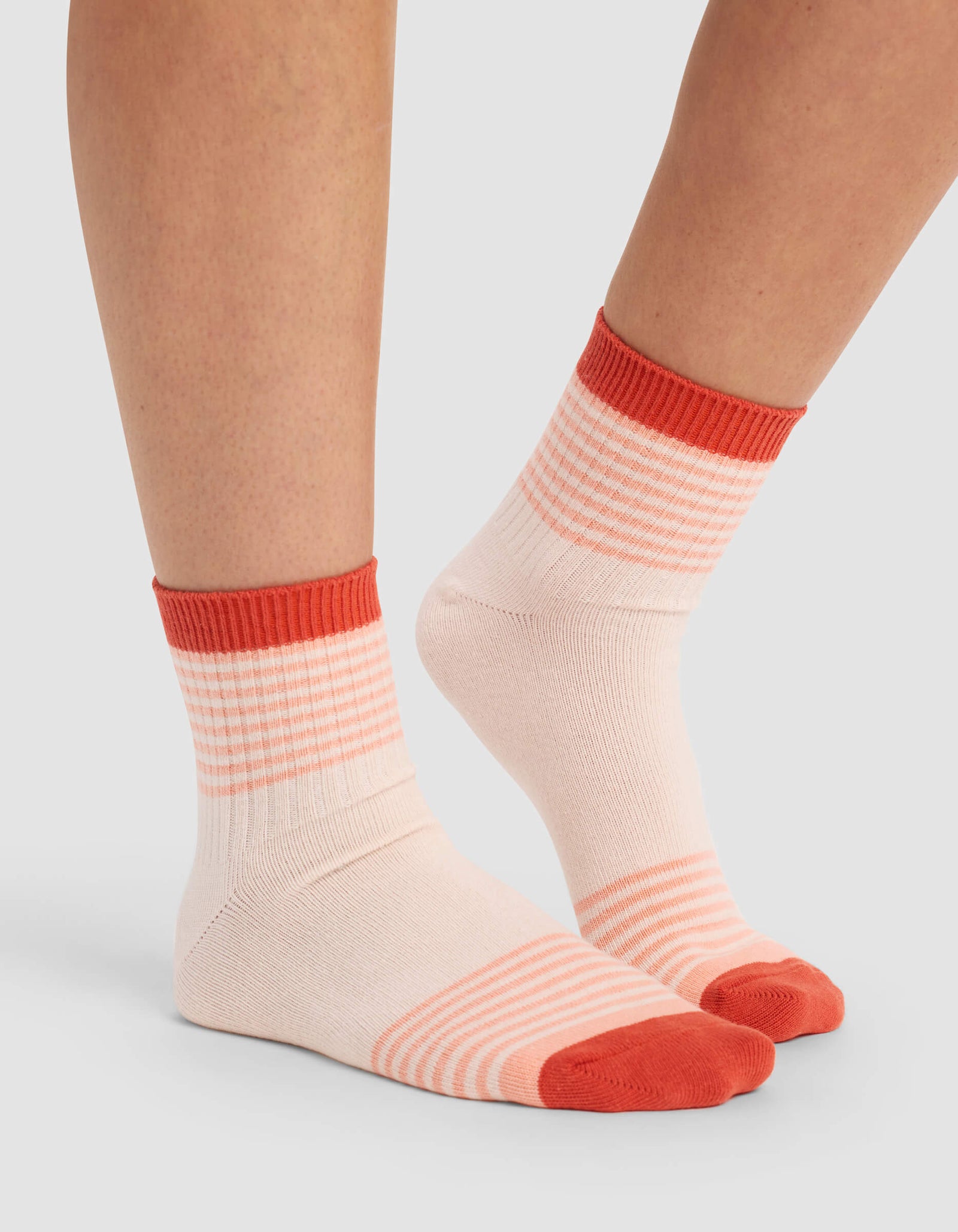A person wears the Cozy Earth Women's Sadie Sock: light pink with orange toes, heels, and cuffs, plus horizontal orange stripes around the ankles and soles. The background is plain and light-colored.