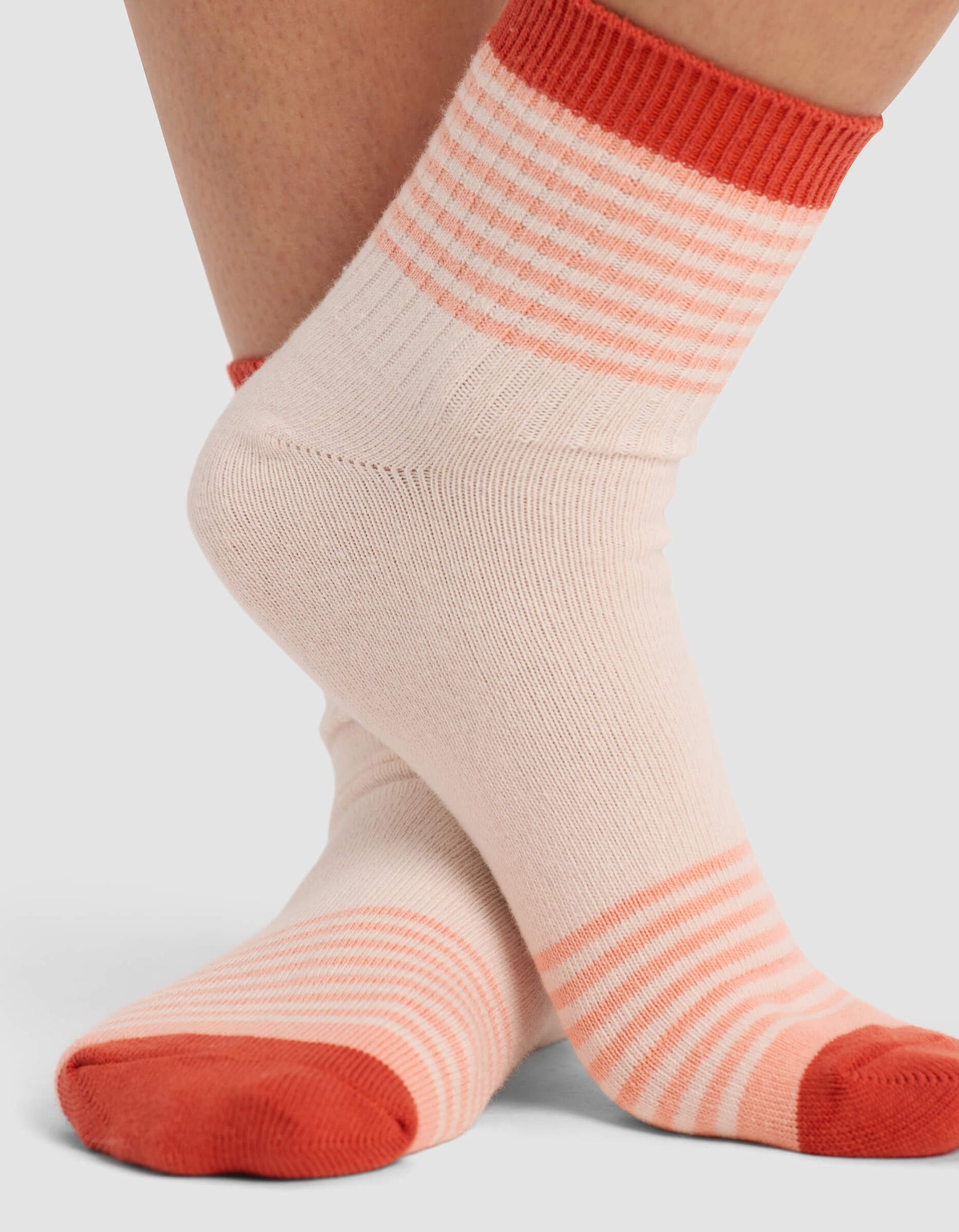 A close-up shows a person with feet crossed at the ankles, wearing Cozy Earth Women's Sadie Socks—light pink with coral horizontal stripes on the toes, heels, and cuffs, plus solid coral accents on the cuff, heel, and toe.