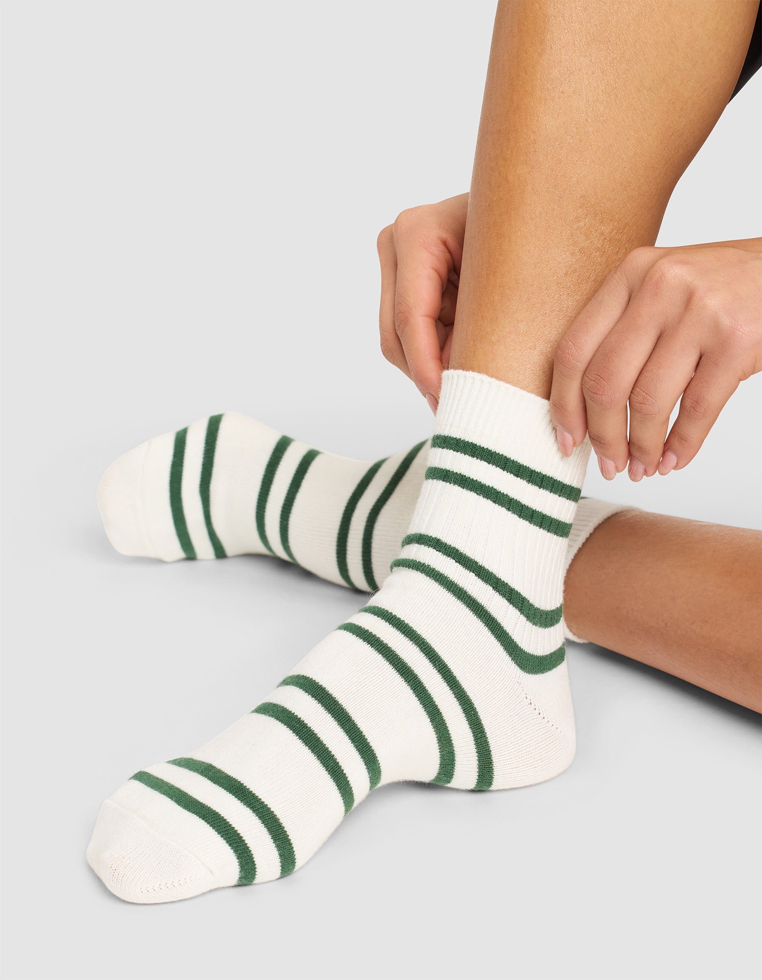 A person wearing Cozy Earth Women's Sadie Socks in white with dark green stripes sits on the floor, adjusting one sock. The photo highlights their legs and feet. 