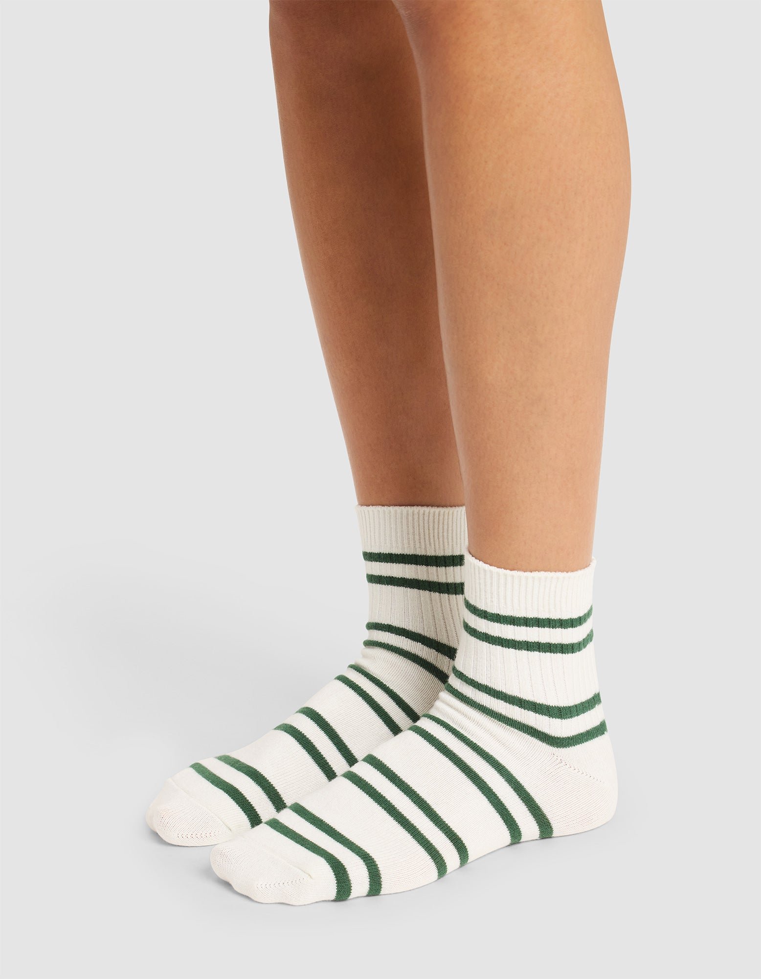 A person wears Cozy Earth Women's Sadie Socks—white with green horizontal stripes reaching just above the ankles—while standing on a light-colored surface. Only their legs and feet are visible. 