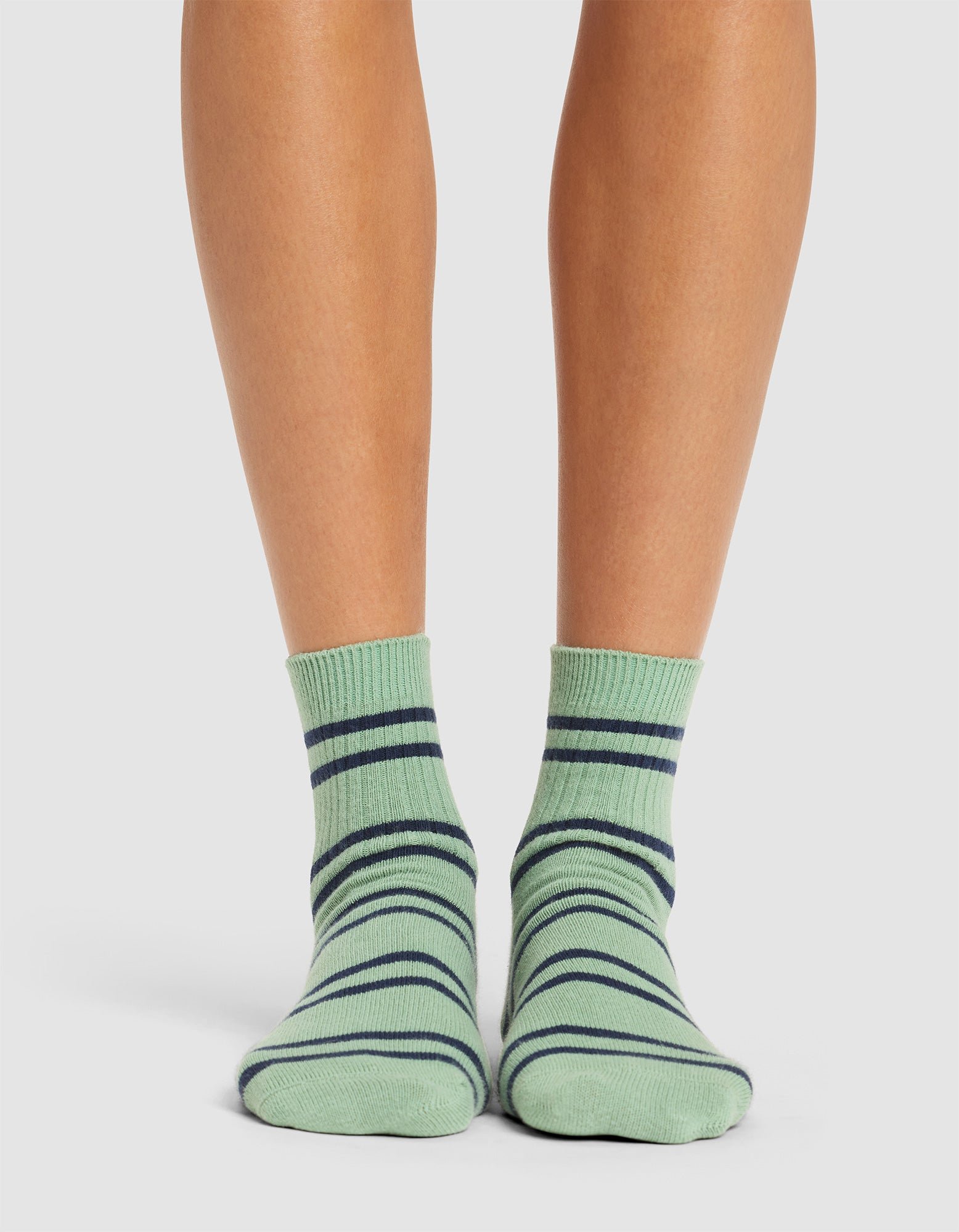 A person is wearing Cozy Earth's Women's Sadie Sock, featuring light green color with dark blue horizontal stripes, standing on a plain, light-colored surface. Only their lower legs and feet are visible. 