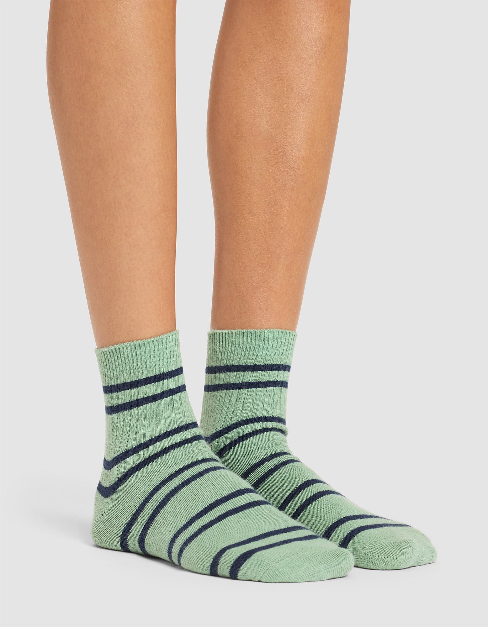 A person wearing Cozy Earth Women's Sadie Socks in light green with dark green horizontal and diagonal stripes, standing on a white background. Only the lower legs and feet are visible. 
