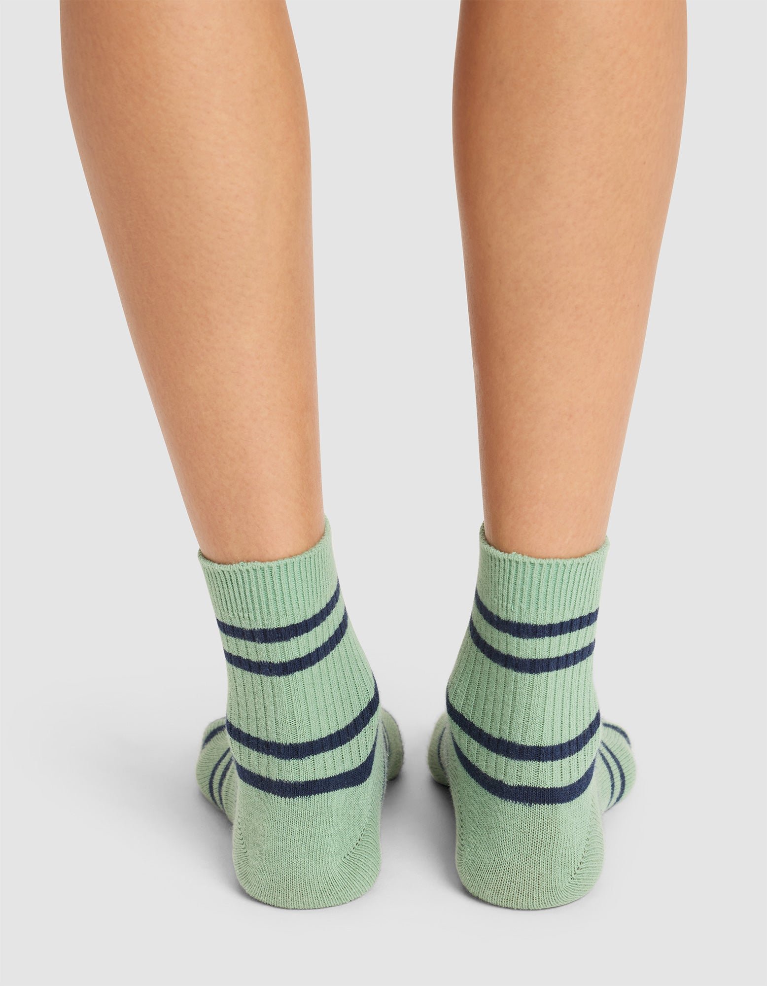 Close-up of a person's lower legs wearing Cozy Earth Women's Sadie Sock in light green with dark blue horizontal stripes, standing on a white background. 