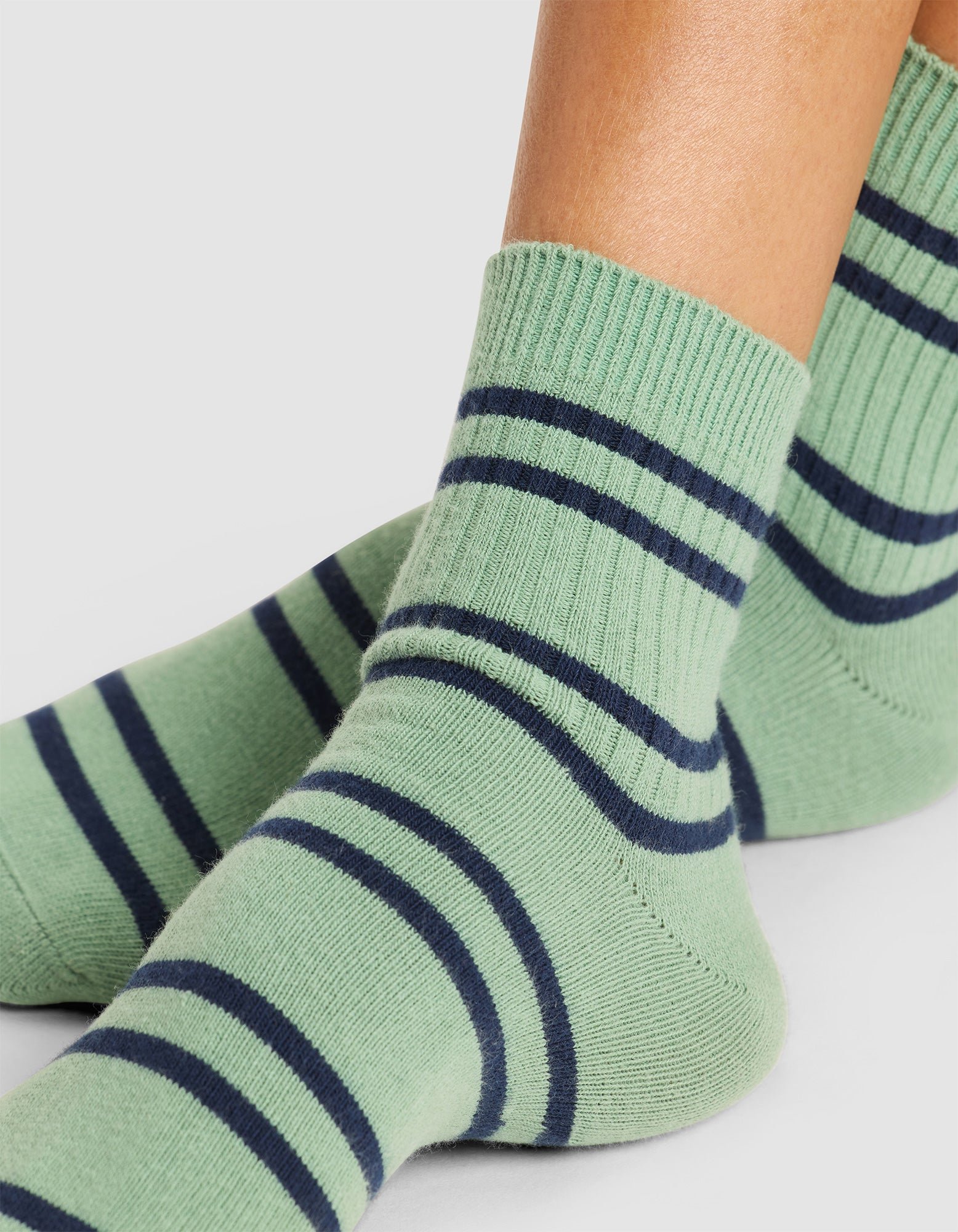Close-up of feet wearing Cozy Earth Women's Sadie Socks in green with dark blue horizontal stripes, displayed on a light gray background. 