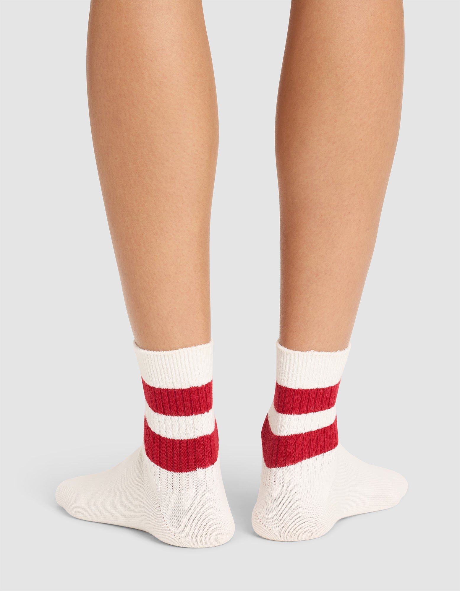 Back view of a person wearing Cozy Earth Women's Sadie Socks—white with two thick red stripes—standing on a light gray surface. Only the lower legs and feet are shown.  