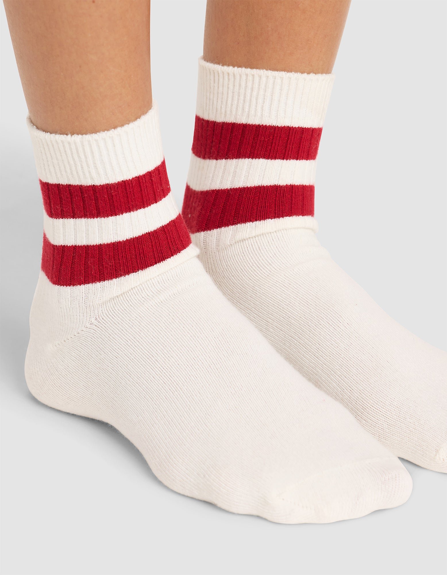 A close-up of feet wearing Cozy Earth Women's Sadie Socks—white crew socks with two bold red stripes near the top—set against a plain, light background. 