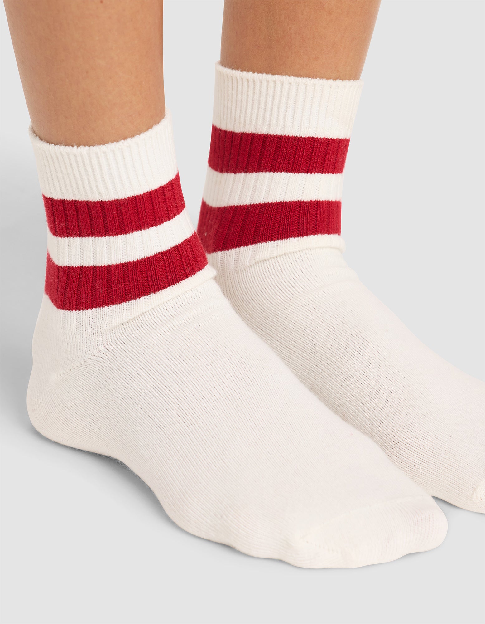 A close-up of feet wearing Cozy Earth Women's Sadie Socks—white crew socks with two bold red stripes near the top—set against a plain, light background. 