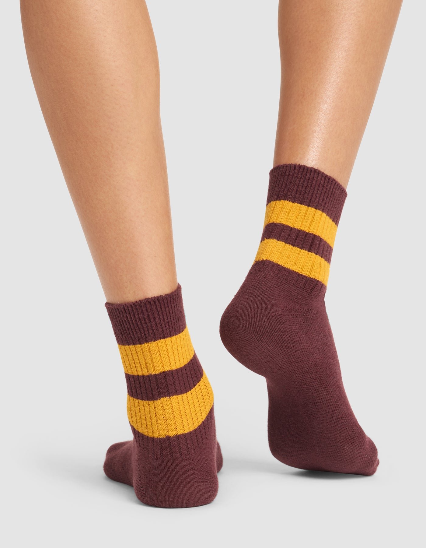 A person wearing Cozy Earth's Women's Sadie Sock—maroon with two thick yellow stripes near the top—stands on a white background, showing only their lower legs and feet. 