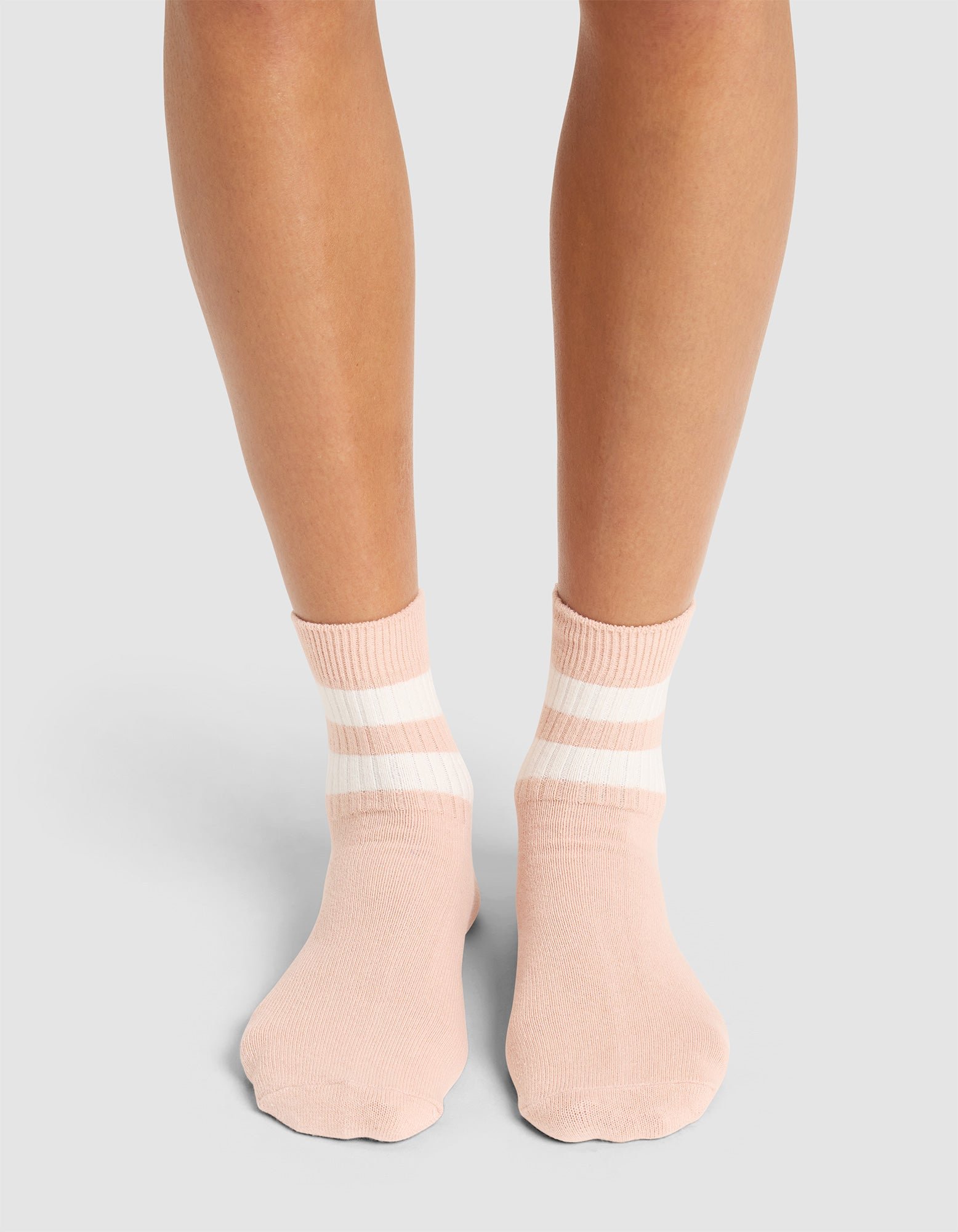 A person stands on a light surface wearing Cozy Earth Women's Sadie Socks in pale pink with two white stripes near the cuffs; only their lower legs and feet are visible. 