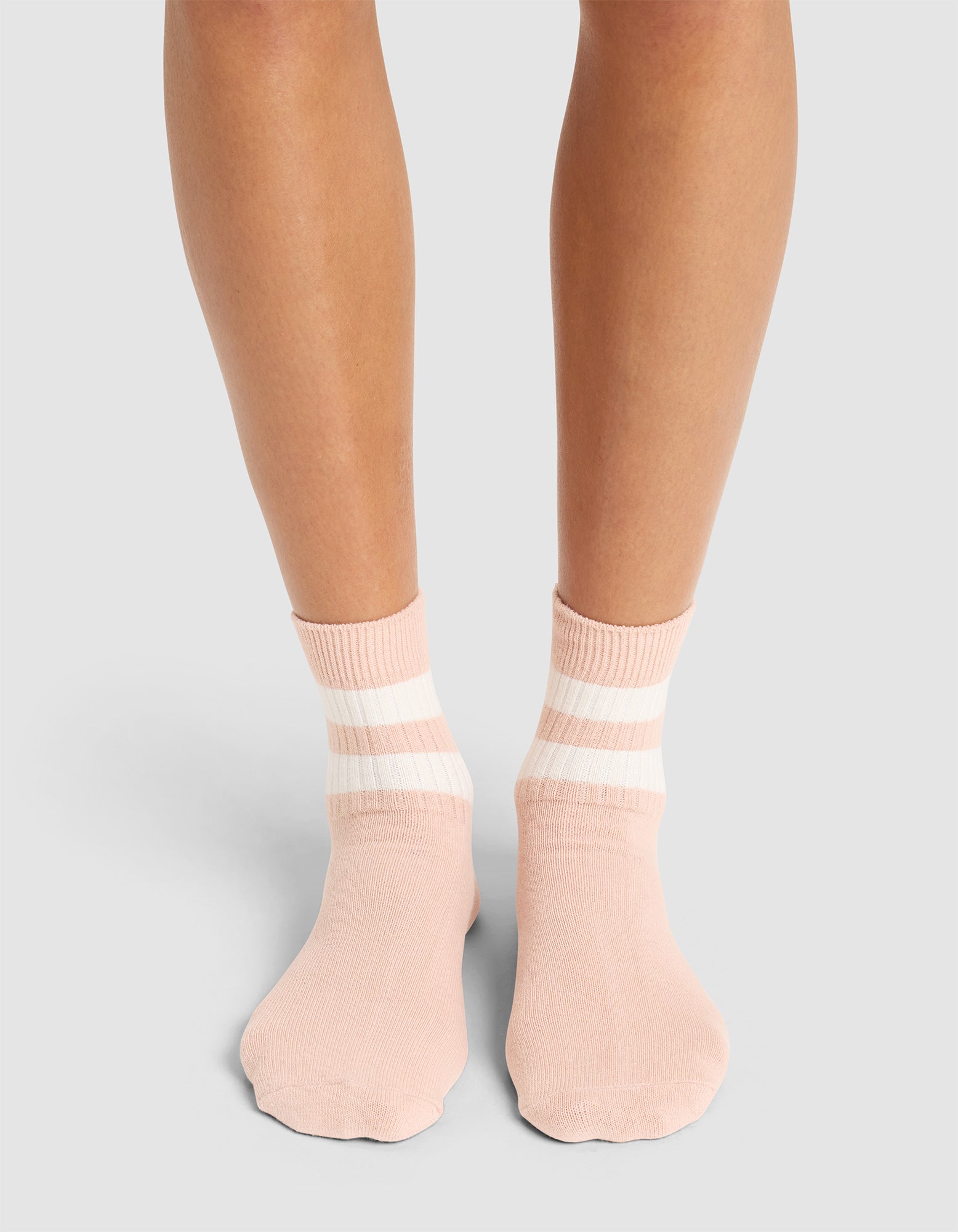 A person stands on a light surface wearing Cozy Earth Women's Sadie Socks in pale pink with two white stripes near the cuffs; only their lower legs and feet are visible. 