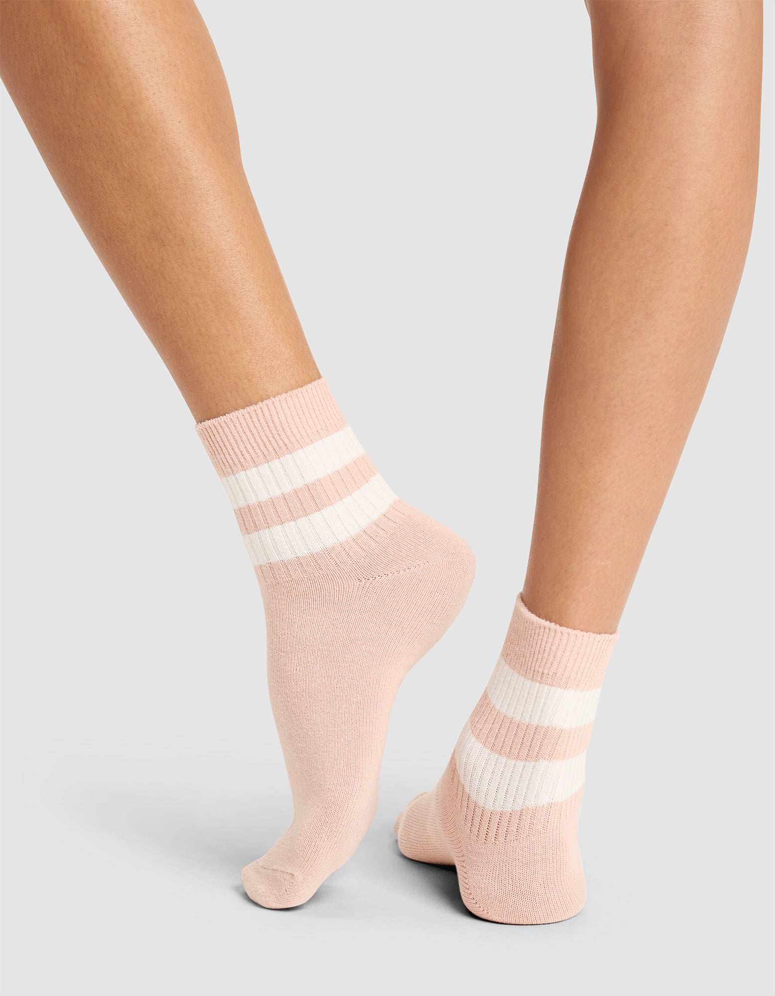 A person stands on tiptoe, showing the back and side of their legs in Cozy Earth Women’s Sadie Socks—pink ankle socks featuring two white horizontal stripes—against a plain light background. 