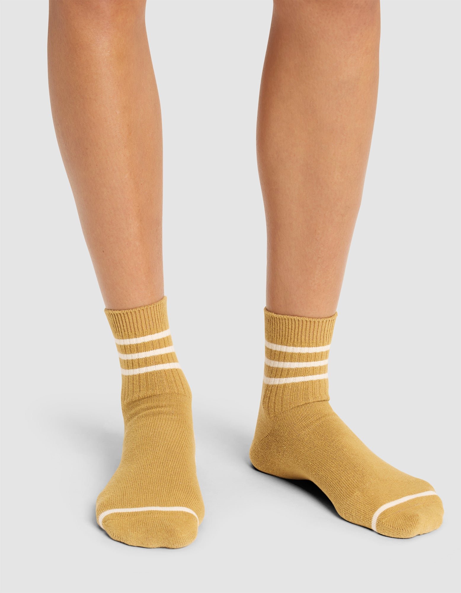 A person wears Cozy Earth Women's Sadie Sock in mustard yellow with two white stripes near the top, standing on a light gray surface. Only their lower legs and feet are visible. 