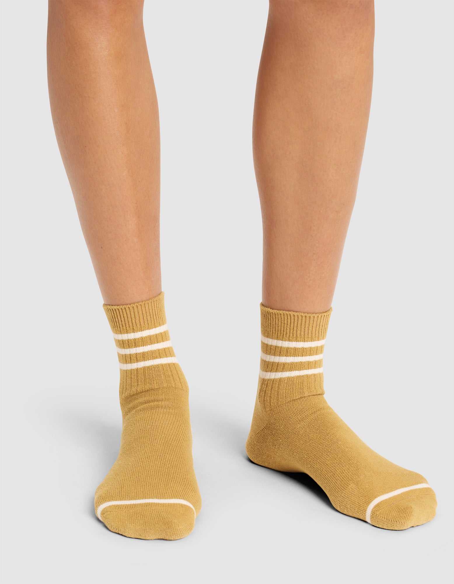 A person wears Cozy Earth Women's Sadie Sock in mustard yellow with two white stripes near the top, standing on a light gray surface. Only their lower legs and feet are visible. 