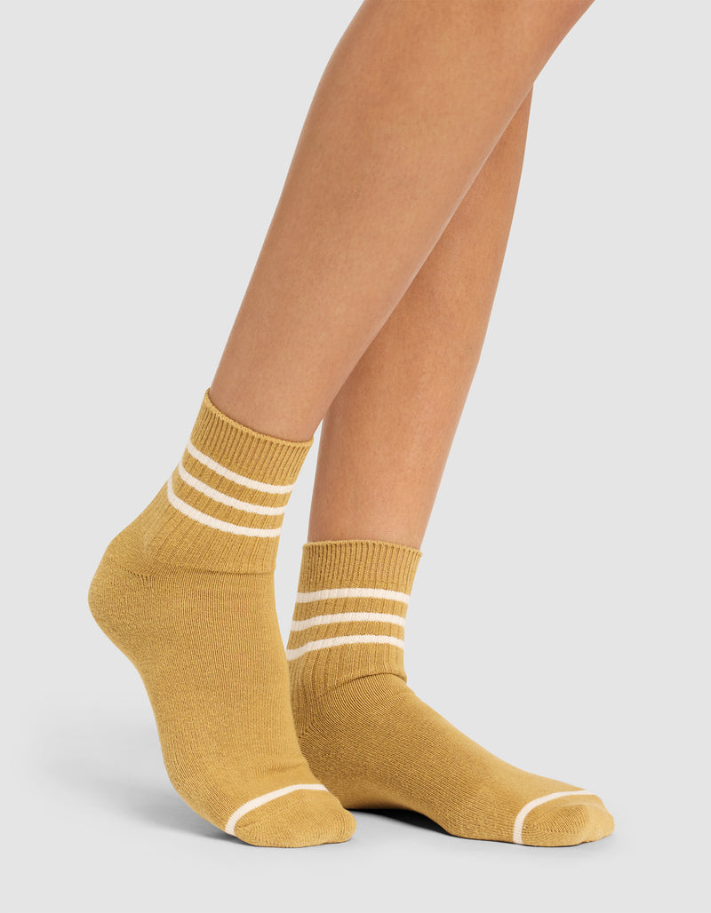 A person wears Cozy Earth Women's Sadie Socks in mustard yellow with white stripes at the ankle and toes, standing against a plain light background.