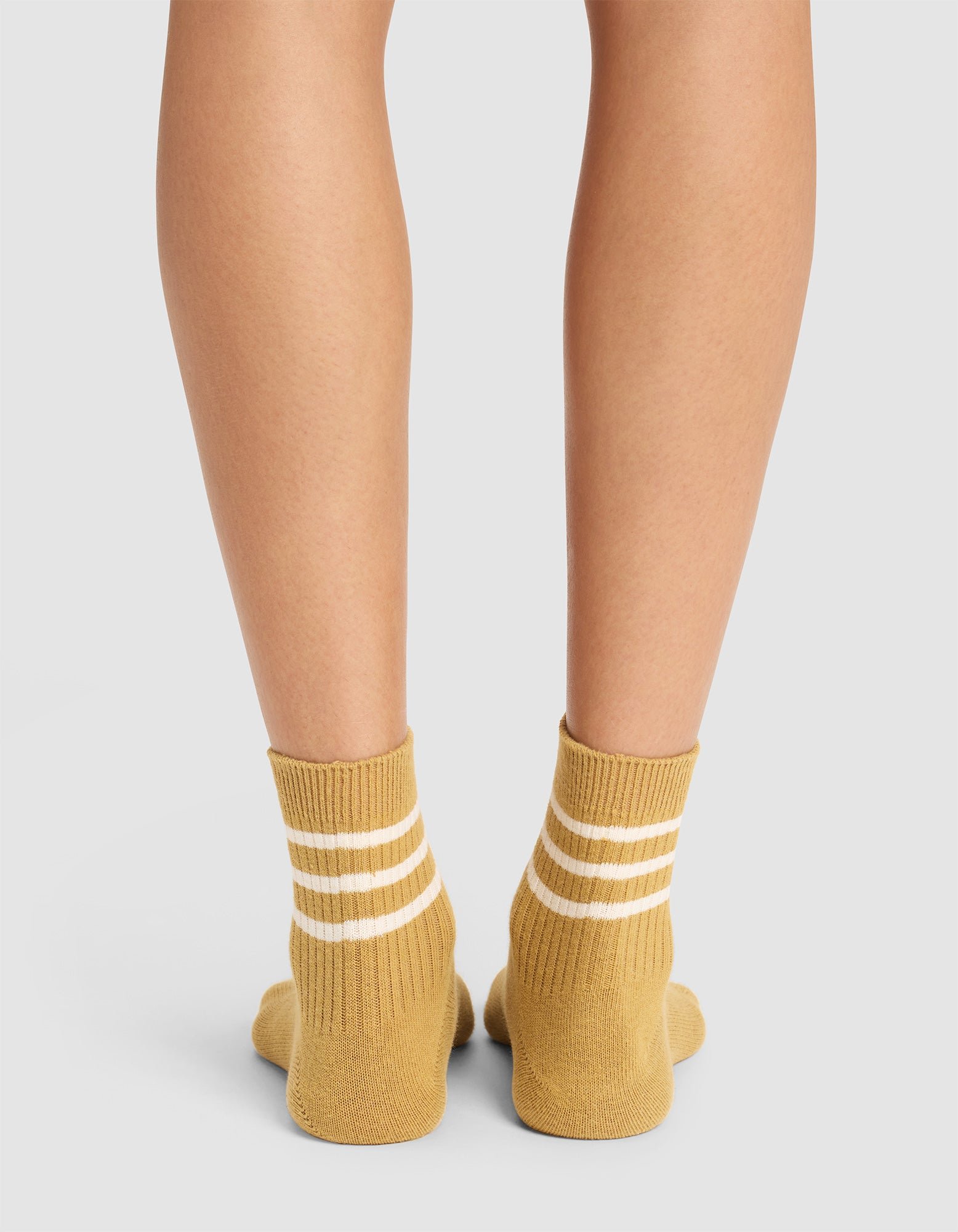 Back view of lower legs and feet wearing Cozy Earth women's Sadie Sock in mustard yellow with two white ankle stripes, shown on a light gray background. 