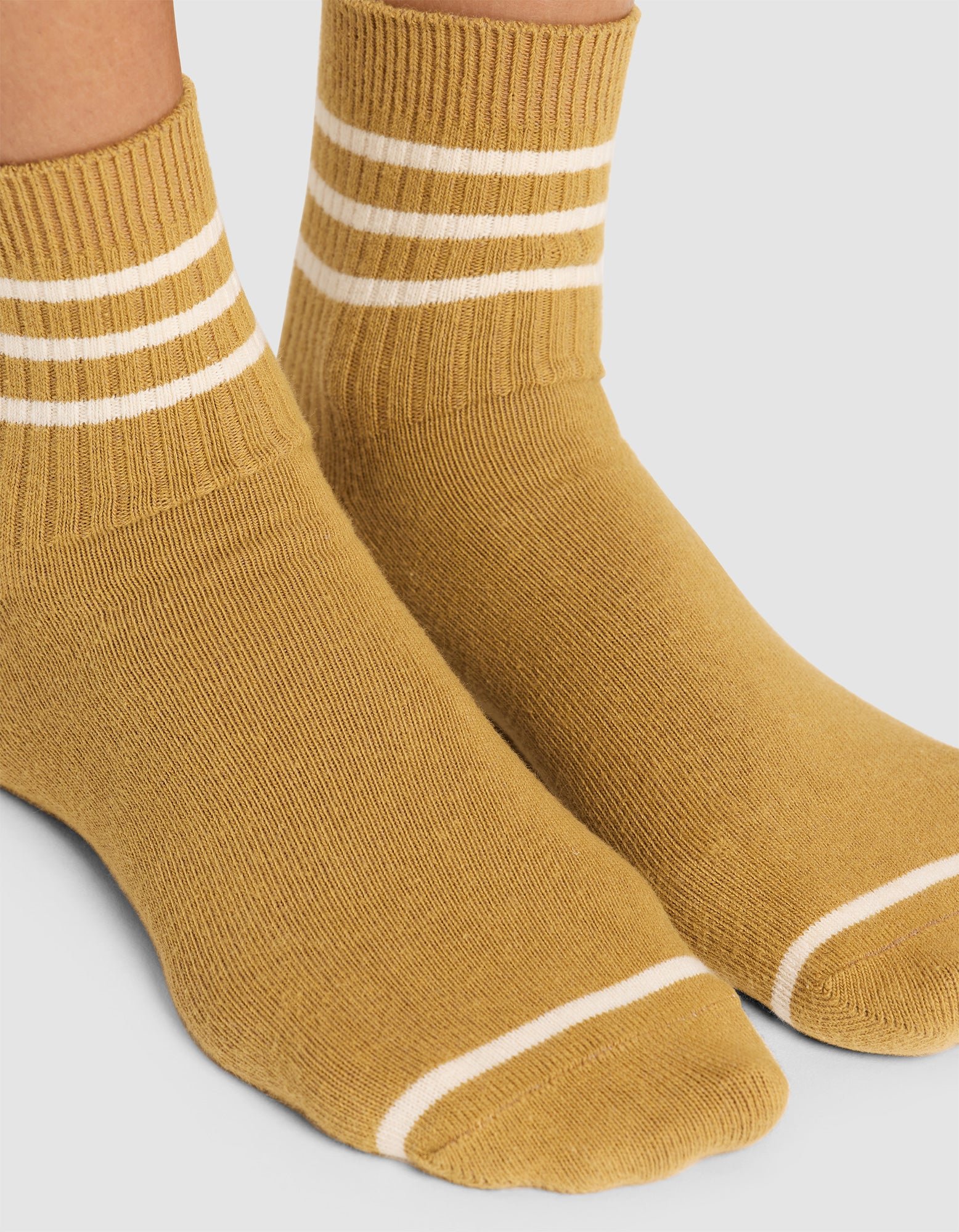 A close-up of feet wearing Cozy Earth Women's Sadie Sock in mustard yellow with white stripes around the cuffs and a single white stripe near the toes, shown against a plain white background. 