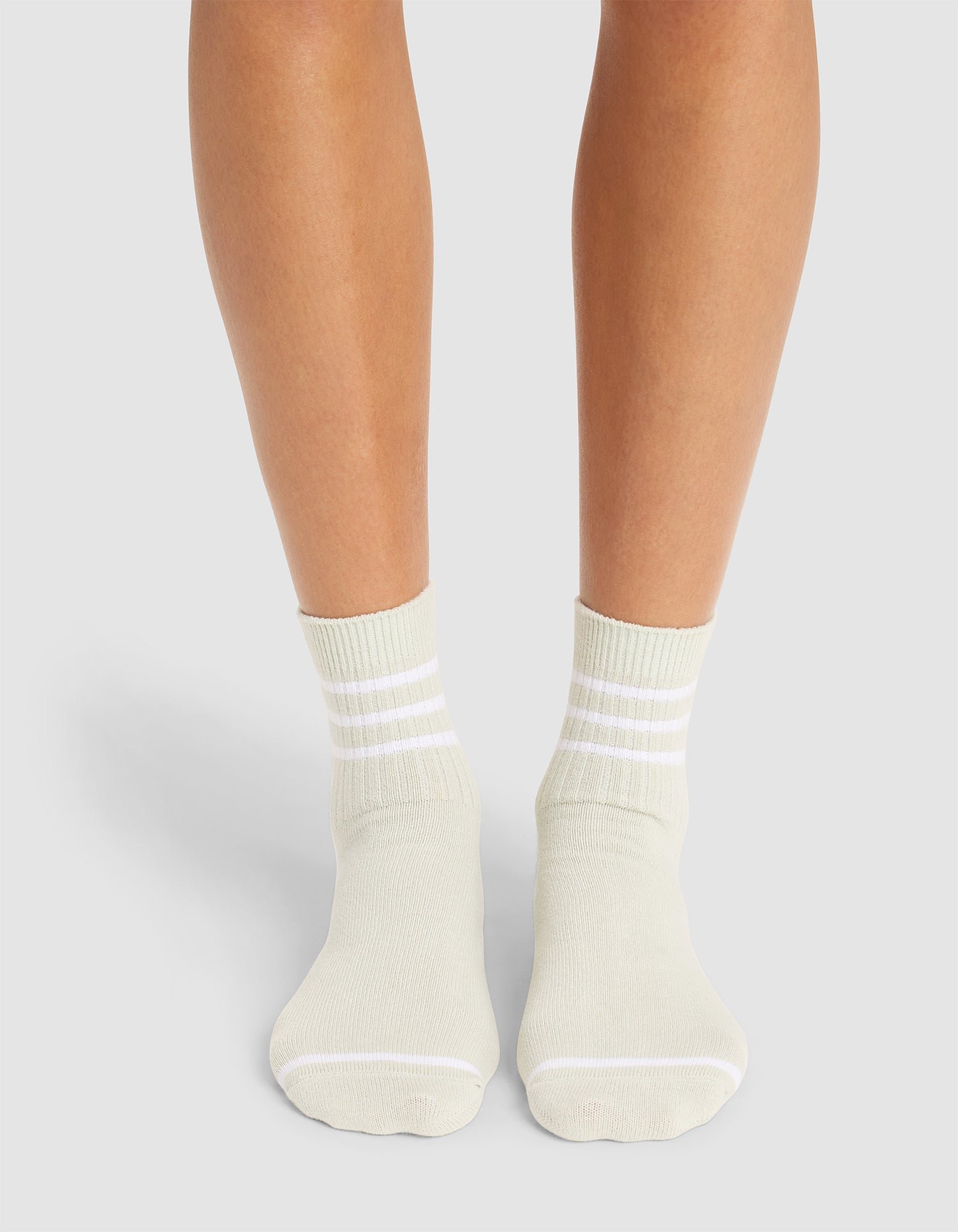 A person wearing Cozy Earth's Women's Sadie Sock in off-white with three white cuff stripes stands on a light grey surface, showing only their lower legs and feet. 