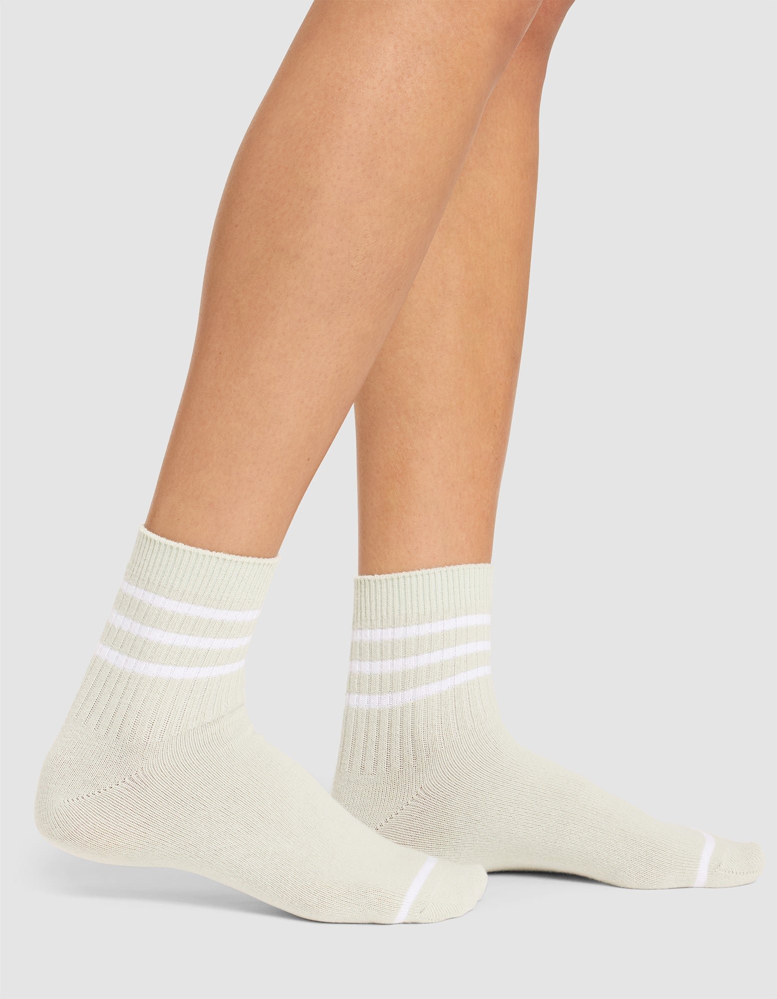 A person is wearing Cozy Earth's Women's Sadie Sock—light-colored ankle socks with three white stripes near the cuffs—while standing on a white background. 