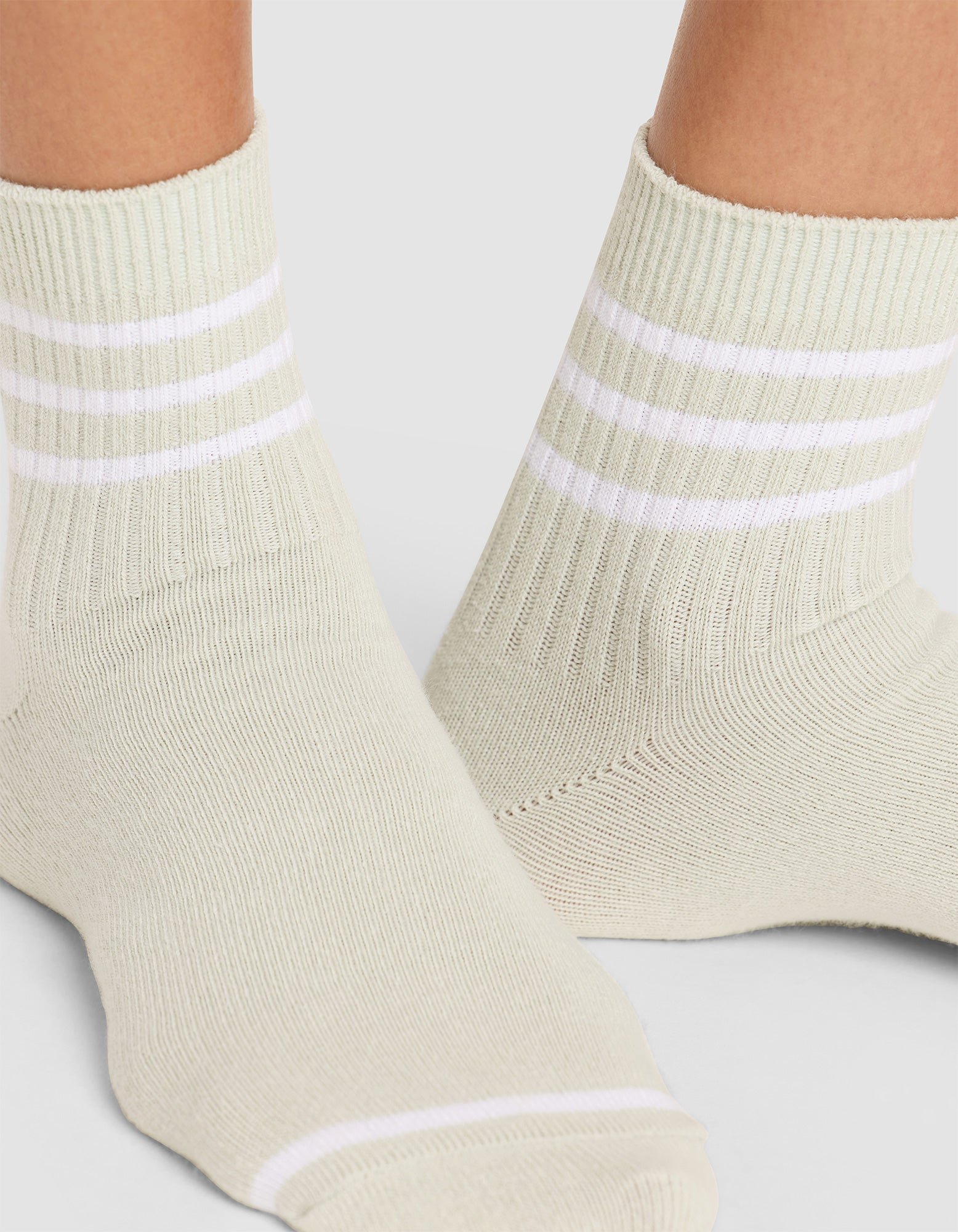 Close-up of two feet wearing Cozy Earth Women's Sadie Socks in light beige with ribbing and three horizontal white stripes at the top, standing on a light gray surface. 