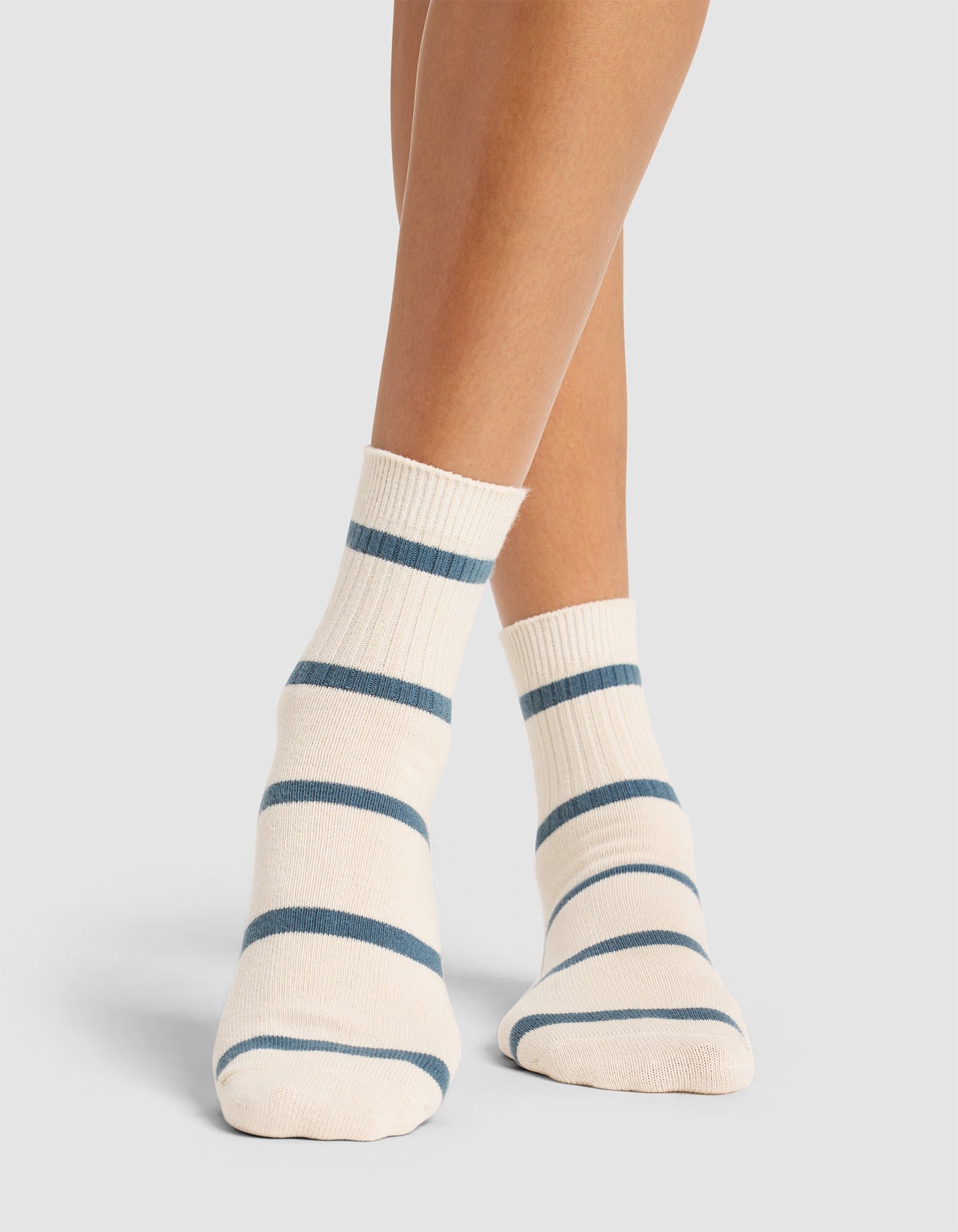 A person stands on a light gray surface wearing Cozy Earth's Women's Sadie Sock—ankle-length white socks with blue stripes—feet slightly apart and legs visible up to mid-calf. 