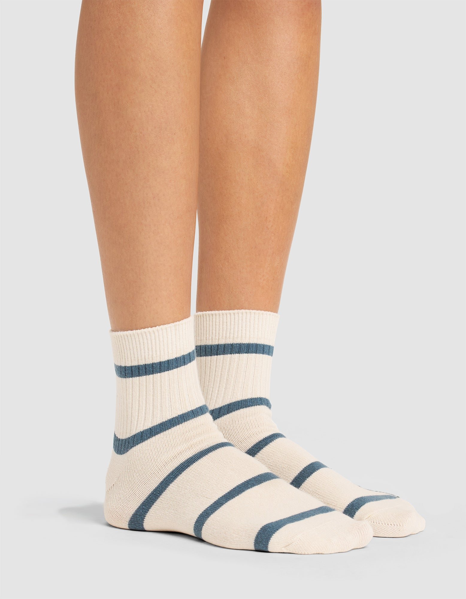 A woman wearing Cozy Earth's Women's Sadie Sock—cream-colored with horizontal blue stripes—stands on a light gray surface. 