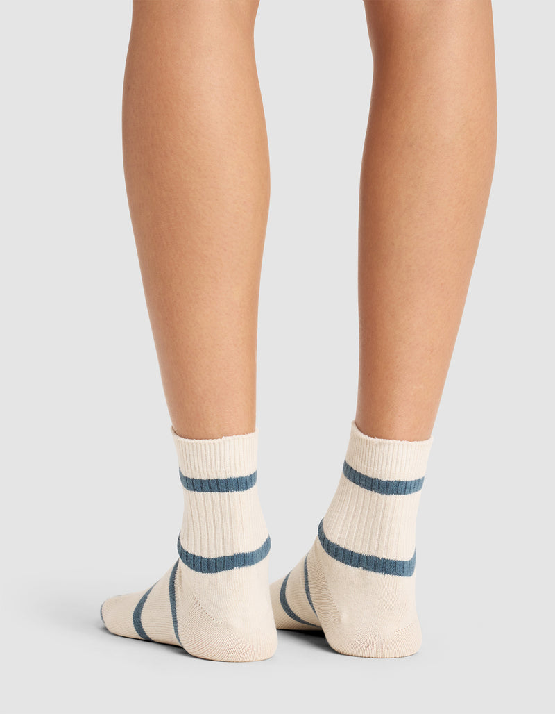 A person standing on a white surface, shown from the calves down, wears Cozy Earth Women's Sadie Socks—cream-colored with two blue horizontal stripes near the top and one stripe by the toes.
