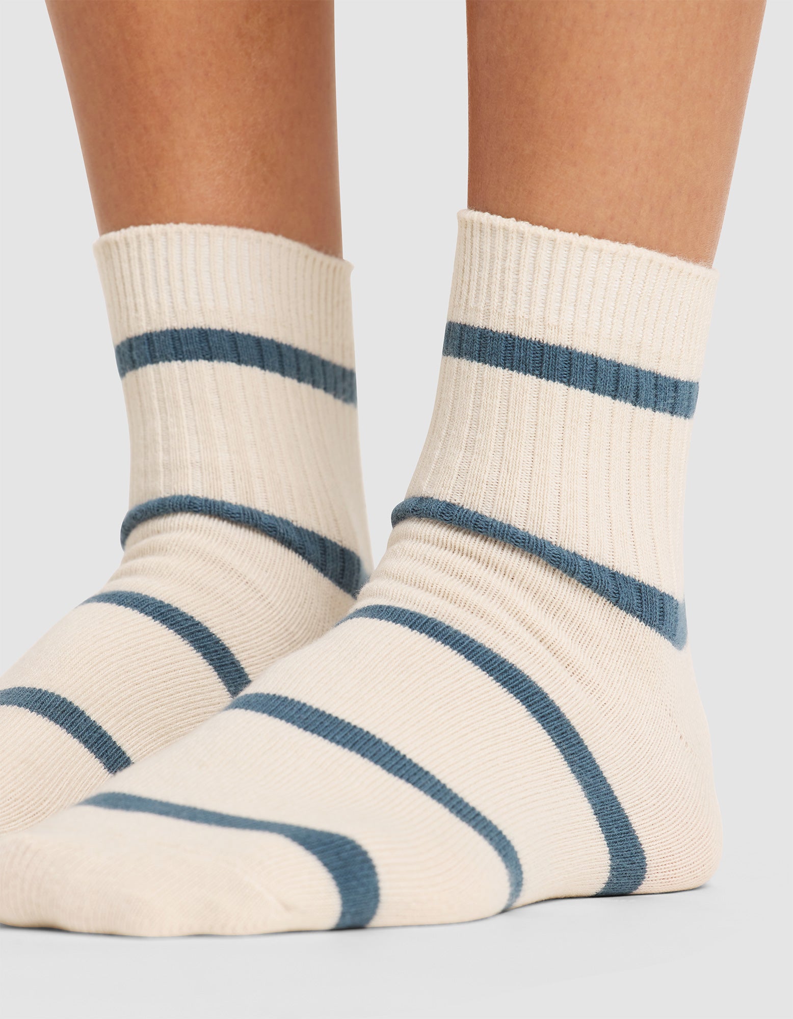Close-up of a person wearing Cozy Earth's Women's Sadie Sock in cream with blue stripes, standing on a light gray surface. Only the lower legs and feet are shown. 