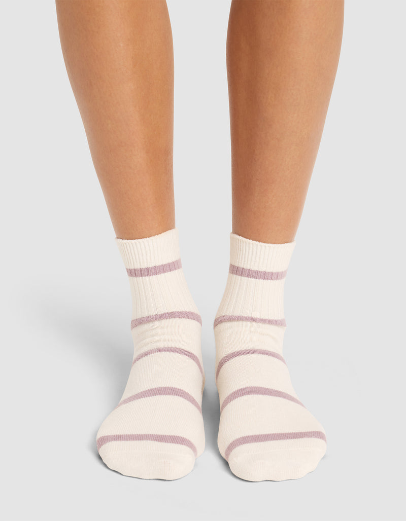 A person stands with feet close together on a light surface, wearing Cozy Earth Women's Sadie Socks—cream-colored with light purple horizontal stripes.