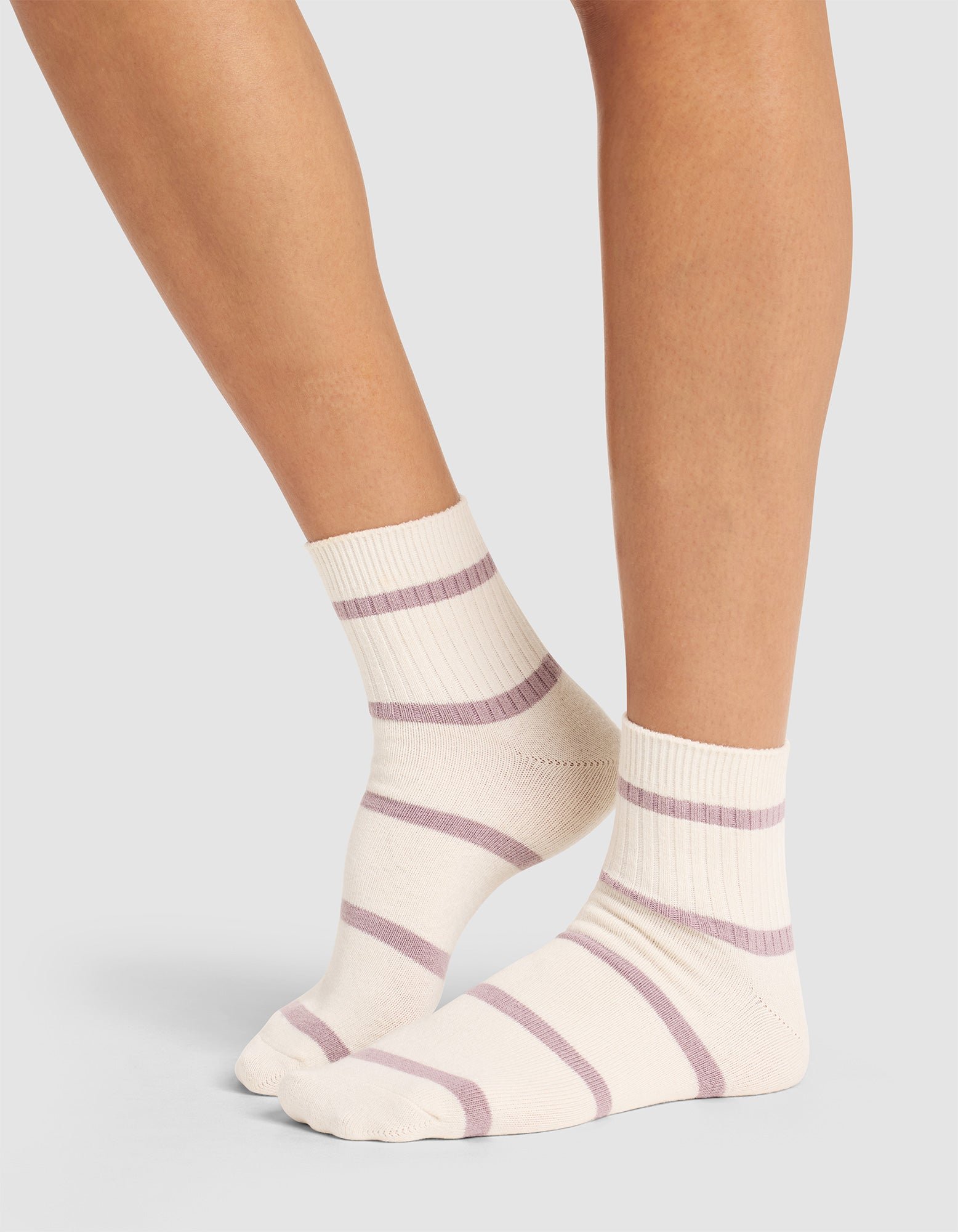 A person stands on tiptoes against a plain light gray background, wearing Cozy Earth's Women's Sadie Sock in cream with light purple horizontal stripes. 