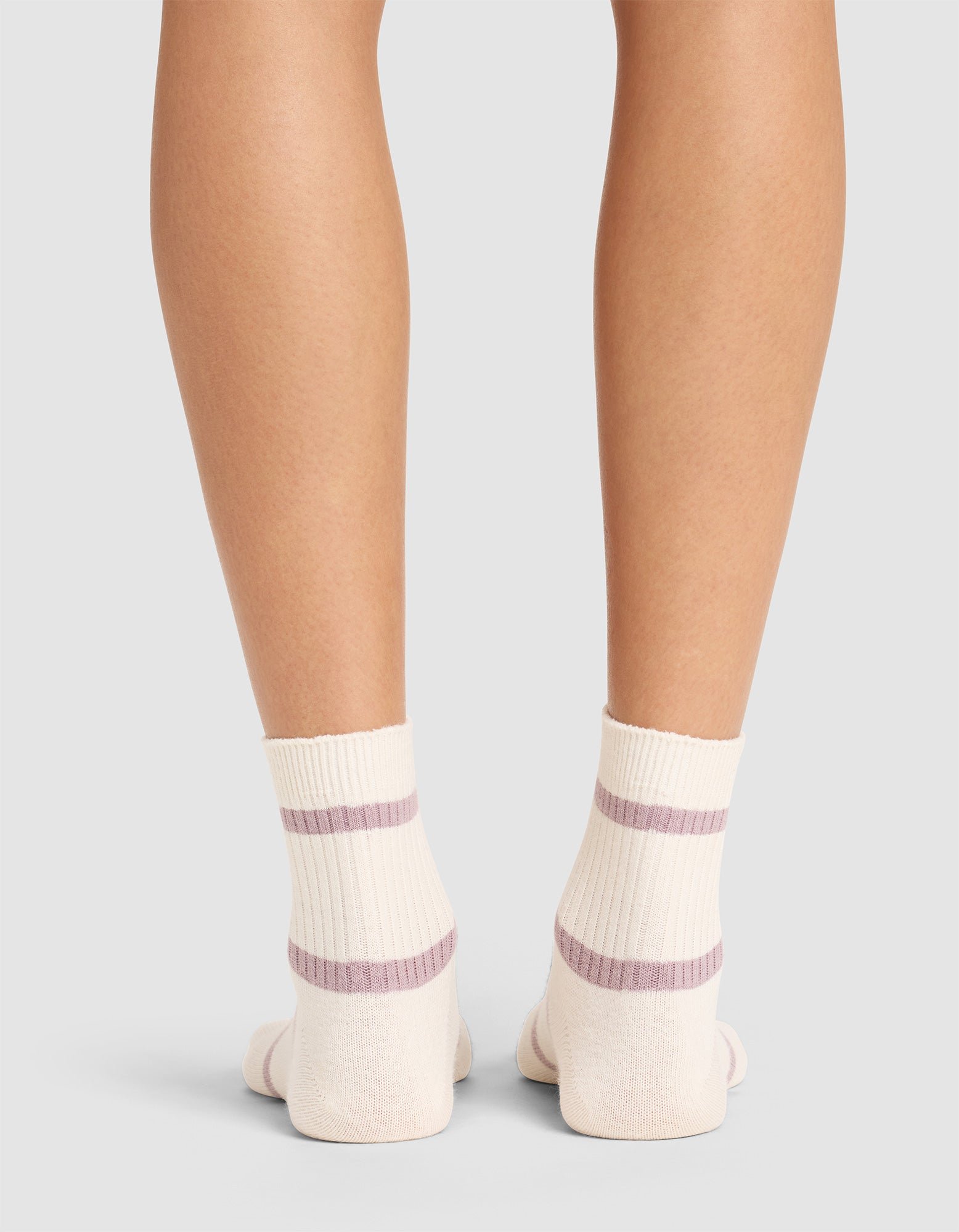 Back view of a person wearing Cozy Earth Women's Sadie Sock in cream with two lavender stripes, standing on a light gray surface. Only the lower legs and feet are visible. 