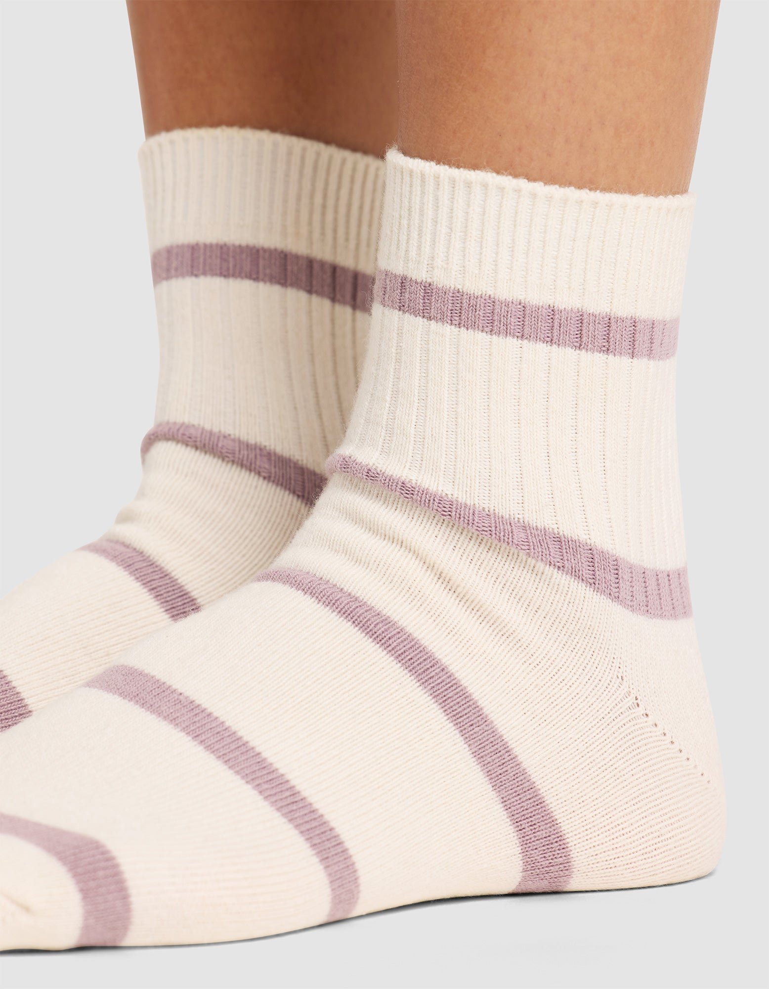 Close-up of two feet wearing Cozy Earth Women's Sadie Socks, featuring cream color with ribbed cuffs and horizontal light purple stripes, standing on a neutral surface. 