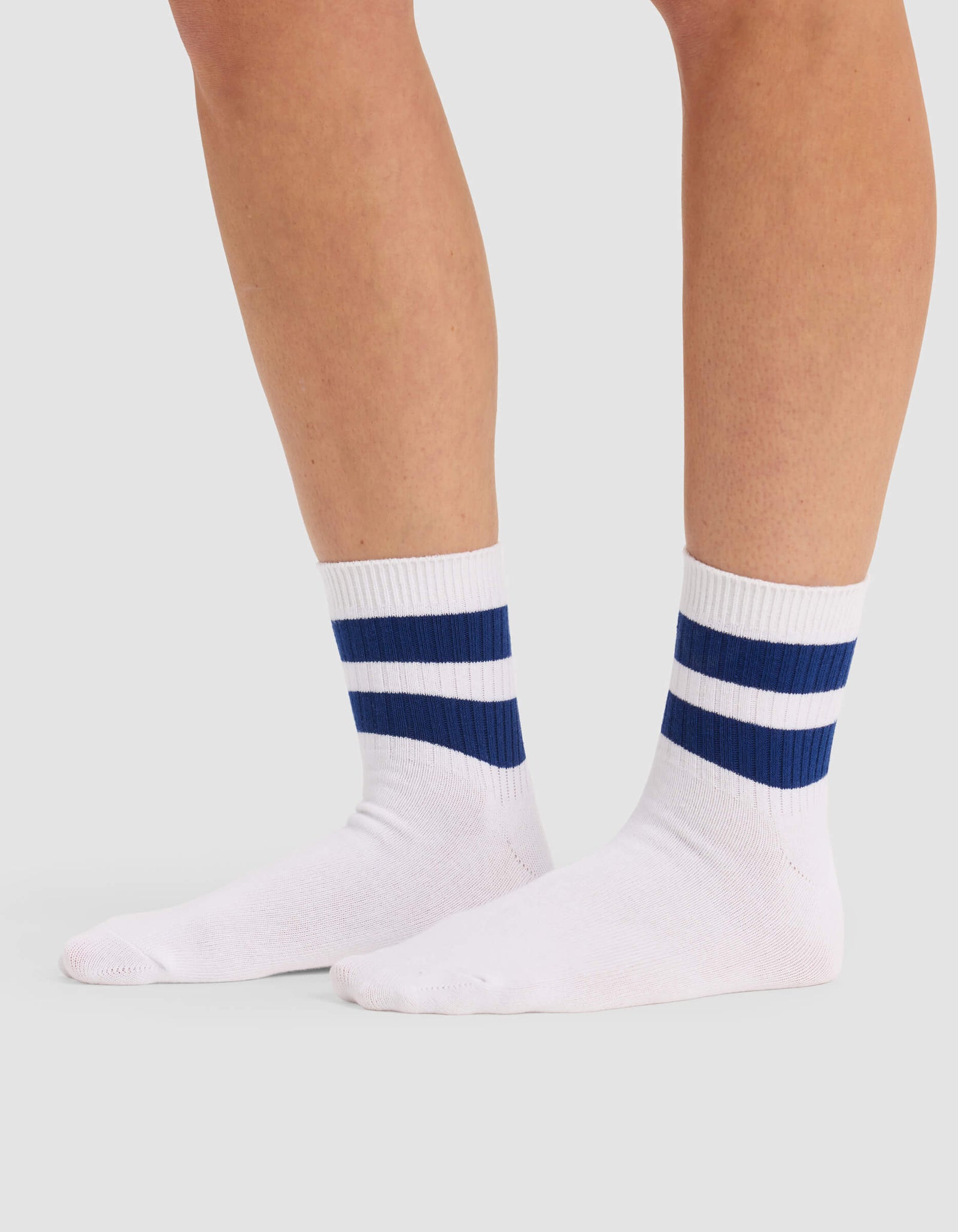 A person stands on a light gray surface wearing Cozy Earth Women's Sadie Socks—white crew socks featuring two horizontal navy blue stripes near the top.