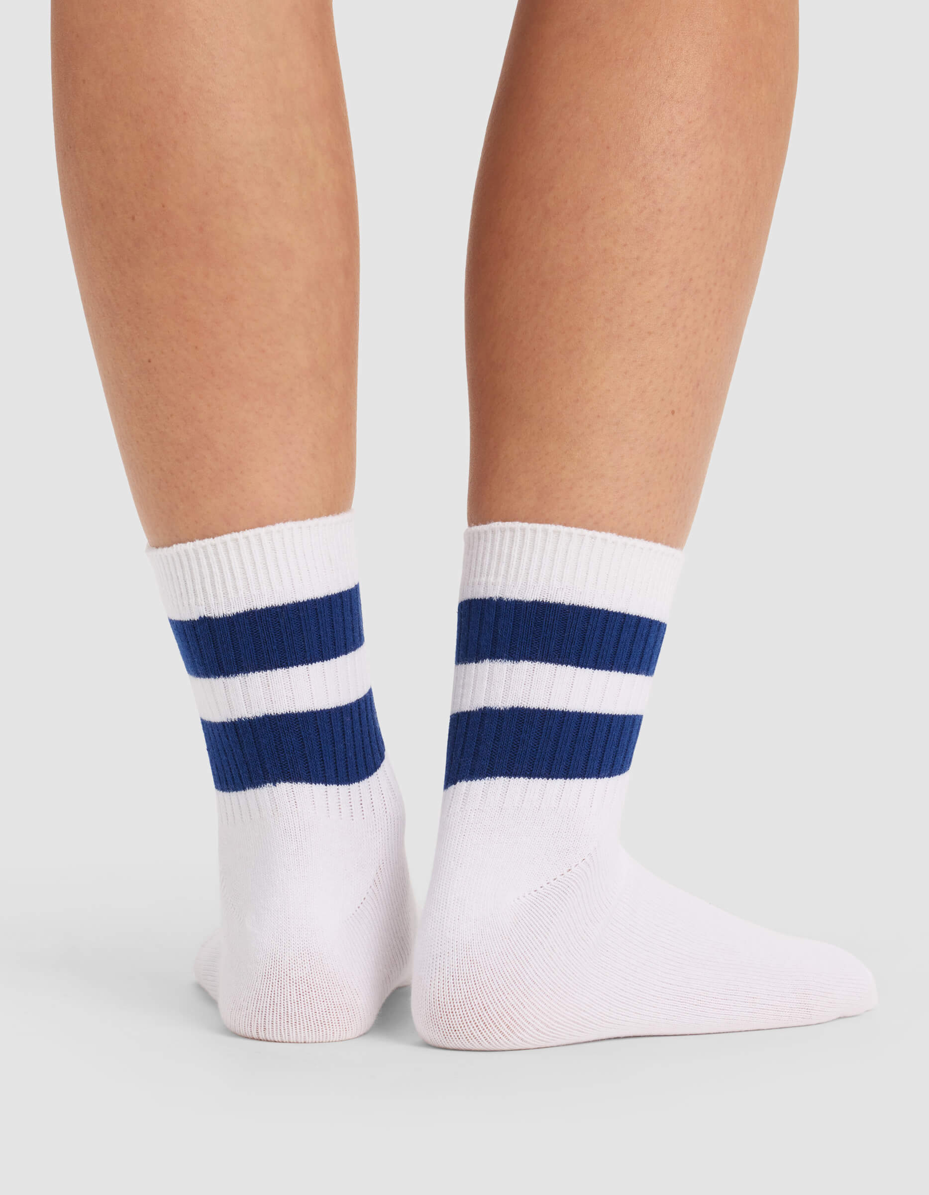A pair of feet wearing Cozy Earth Women's Sadie Socks—white crew socks with two thick blue stripes near the top—standing against a light gray background.