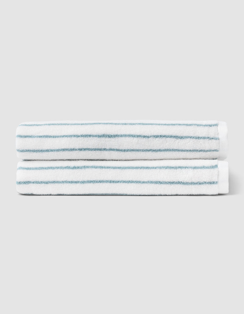 Two Striped Luxe Bath Towels by Cozy Earth, featuring thin blue stripes on white, are stacked together against a plain light gray background.